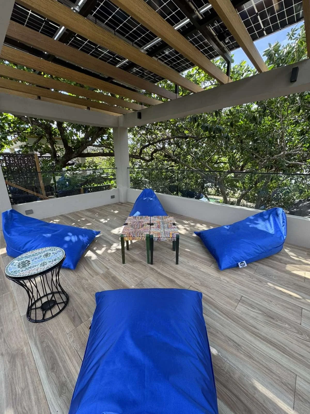 Outdoor balcony with wooden pergola, blue bean bags, a small table, and surrounded by green trees.