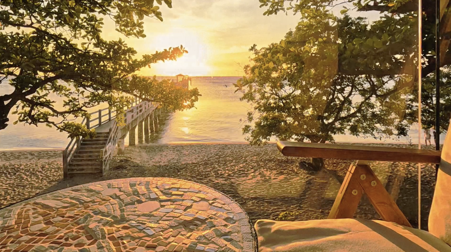 Sunset view over a beach with a jetty, trees, and a tiled table in the foreground.