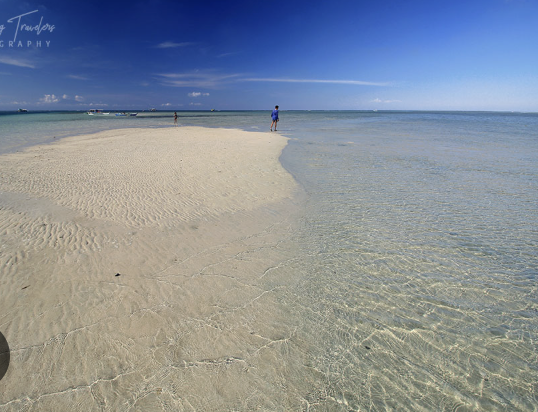 A sandy beach with calm, clear water and a bright blue sky. Two people are walking along the shoreline.