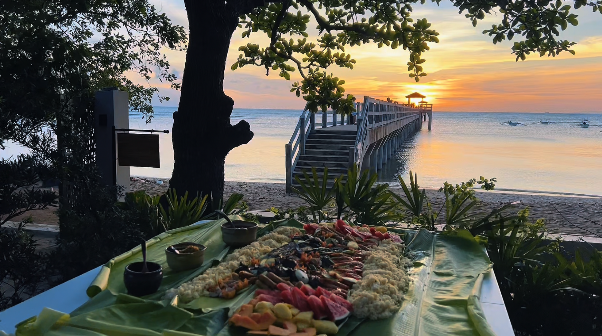 A tropical sunset view over a beach with boats and a pier, with a table of assorted food items in the foreground.