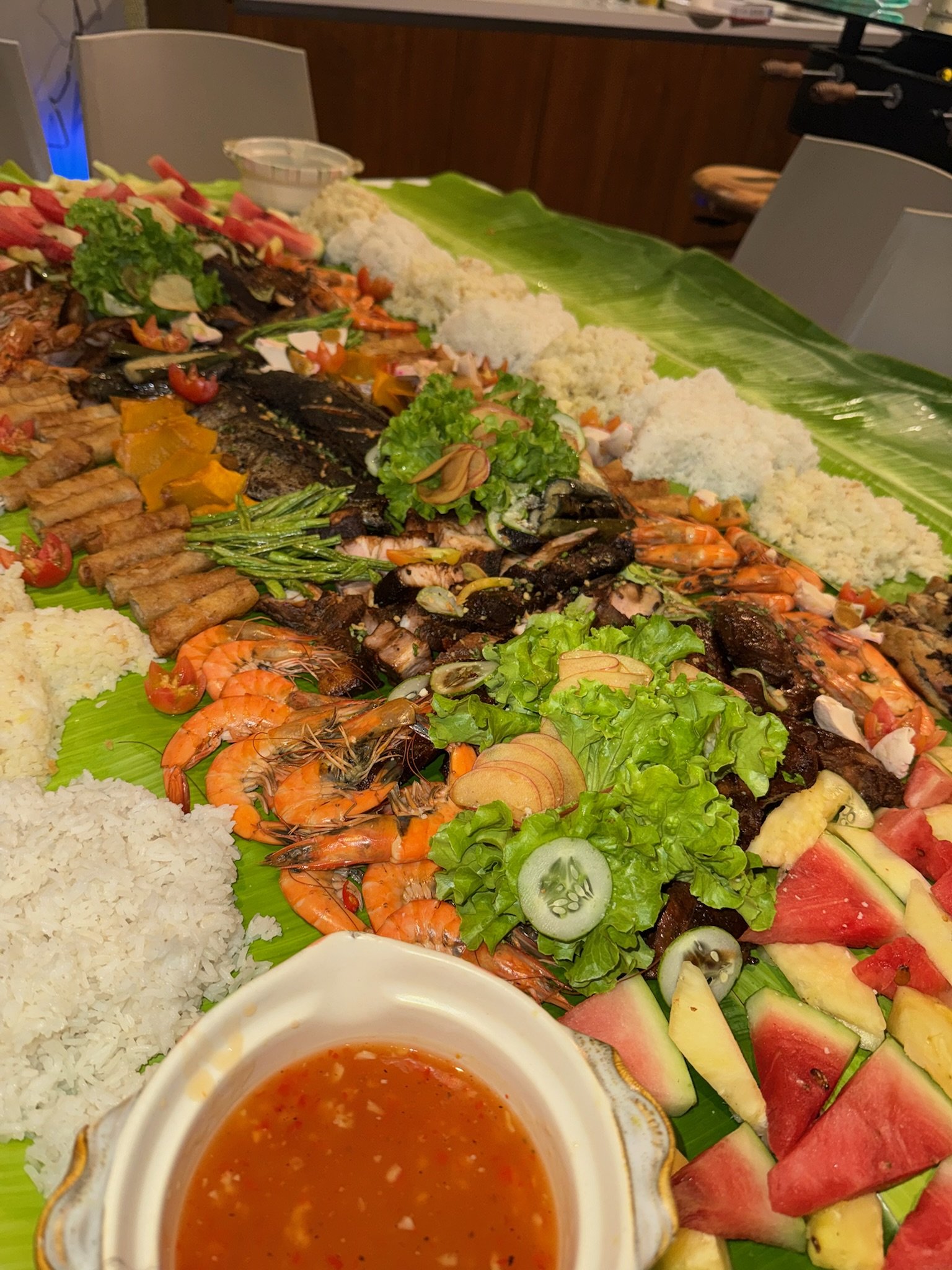 Banana leaf serving tray with an assortment of seafood, rice, vegetables, sliced fruits, and dipping sauce.