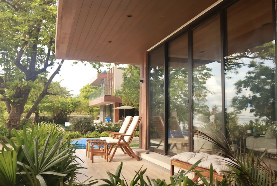 A backyard patio area with wooden outdoor chairs and a bench, surrounded by greenery and bushes, with a large glass sliding door leading into a modern house.