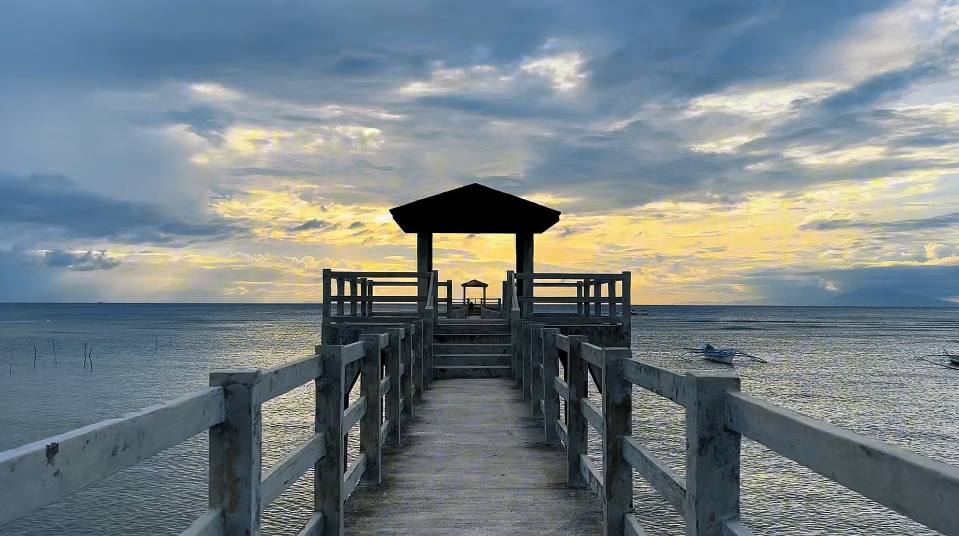 A wooden pier extending over the water toward a small shelter at sunset, with boats floating nearby and a sky filled with clouds and fading sunlight.