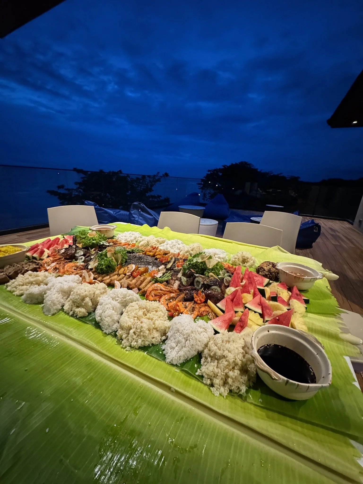 Large platter of assorted tropical fruits, meat and seafood on a banana leaf (Boodle Fight), with dipping sauces, set outdoors during dusk with a view of the ocean and sky.