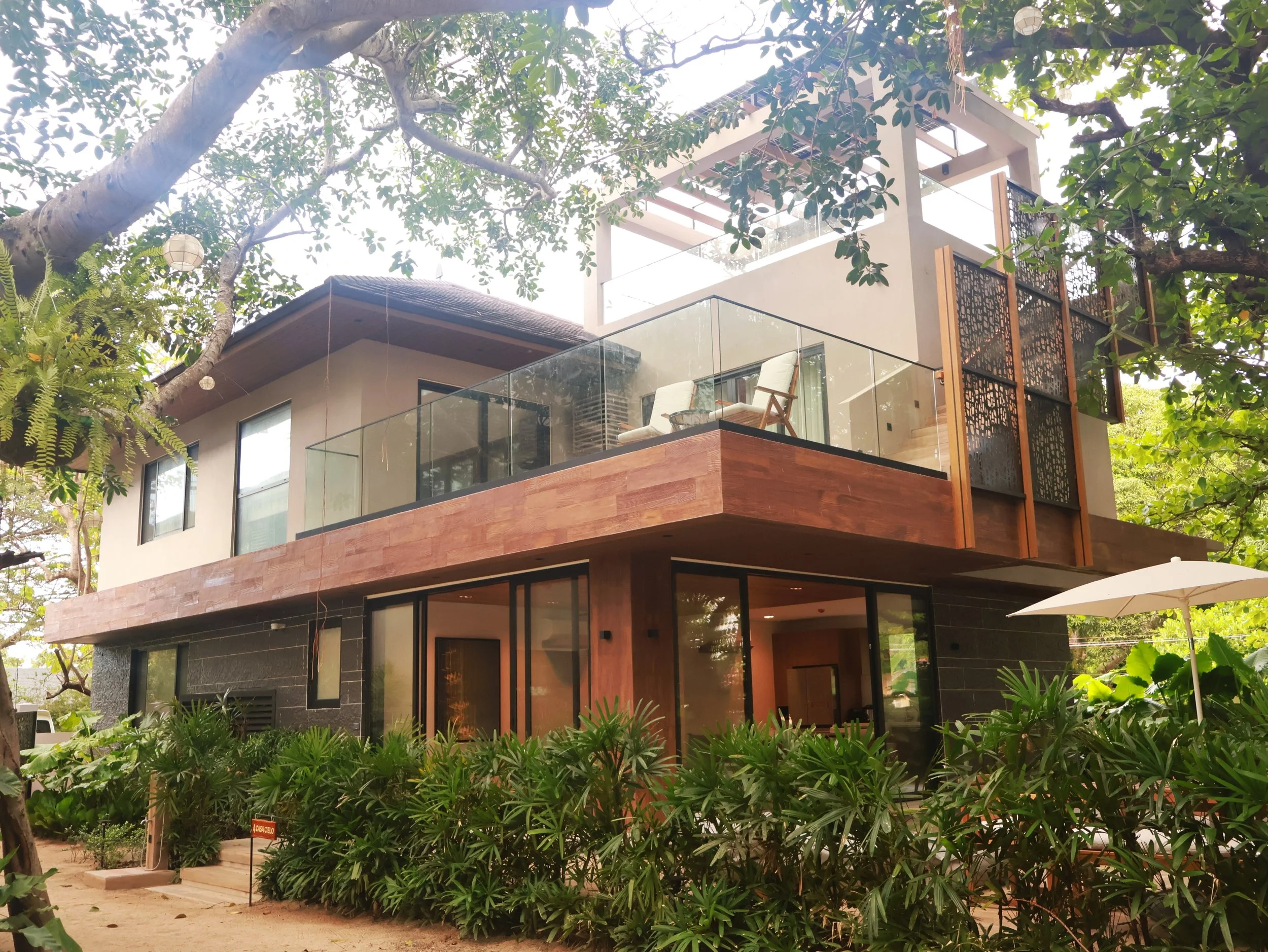 Modern multilevel house surrounded by green trees and plants, featuring glass railings, wooden accents, and large windows, with outdoor seating and an umbrella.