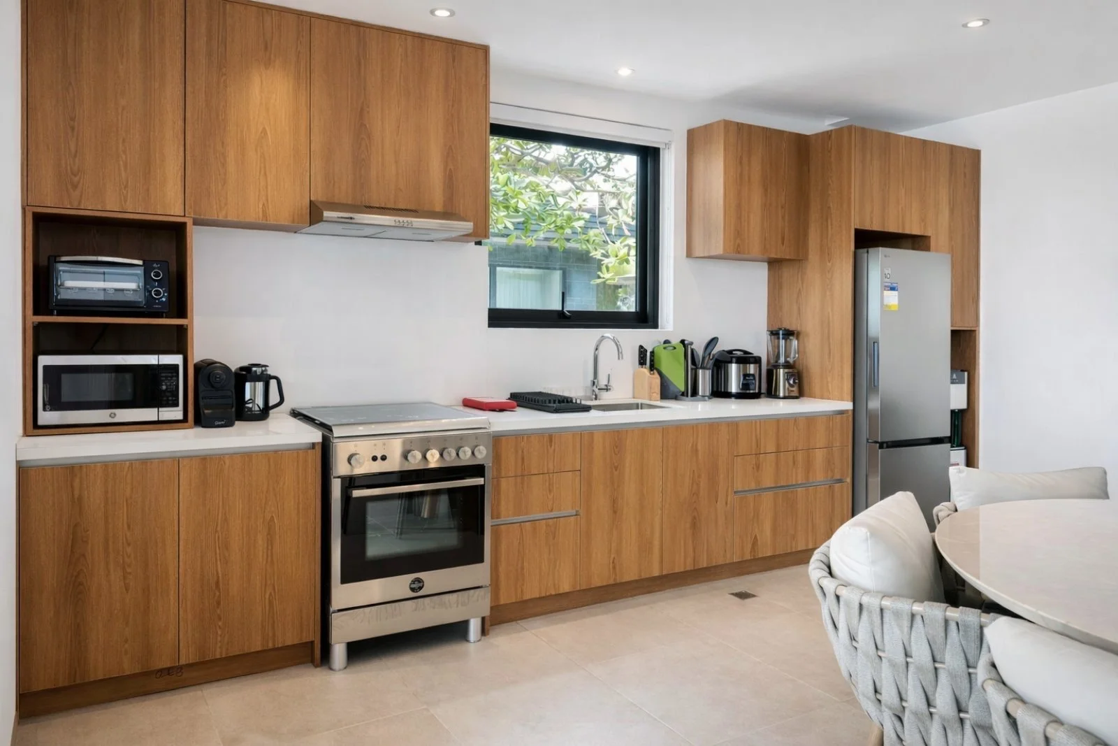 Modern kitchen with wooden cabinets, a window above the sink, stainless steel appliances, including an oven, refrigerator, microwave, and toaster. Small appliances on the countertop include a coffee maker and blender. Part of a round dining table with chairs is visible in the foreground.