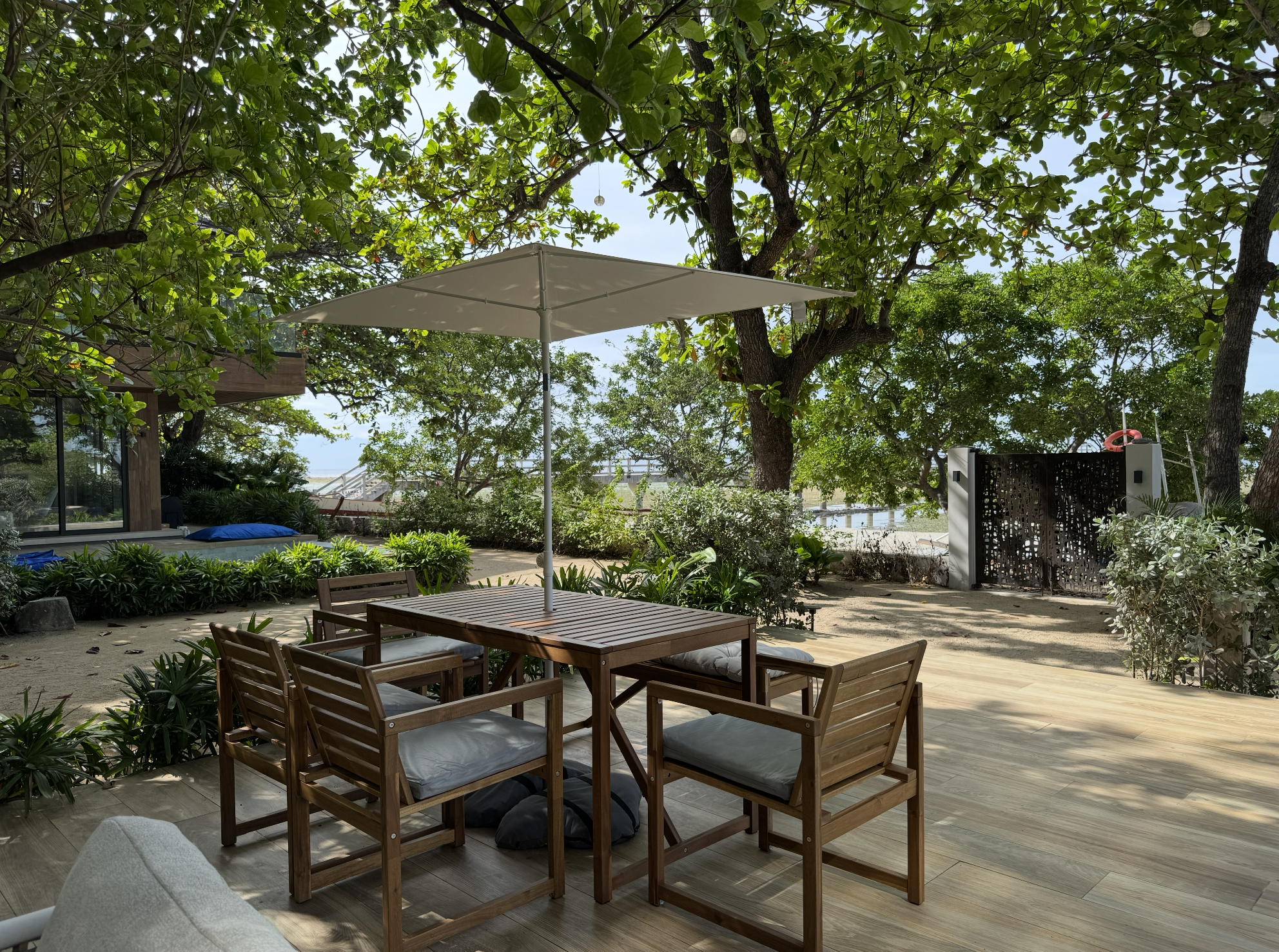 Outdoor patio with wooden table and chairs, a white umbrella, surrounded by trees and greenery, with a sandy ground and some plants.