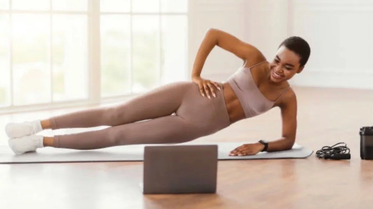Woman exercising on yoga mat at home, doing side plank while watching a laptop, in a bright room with large windows.
