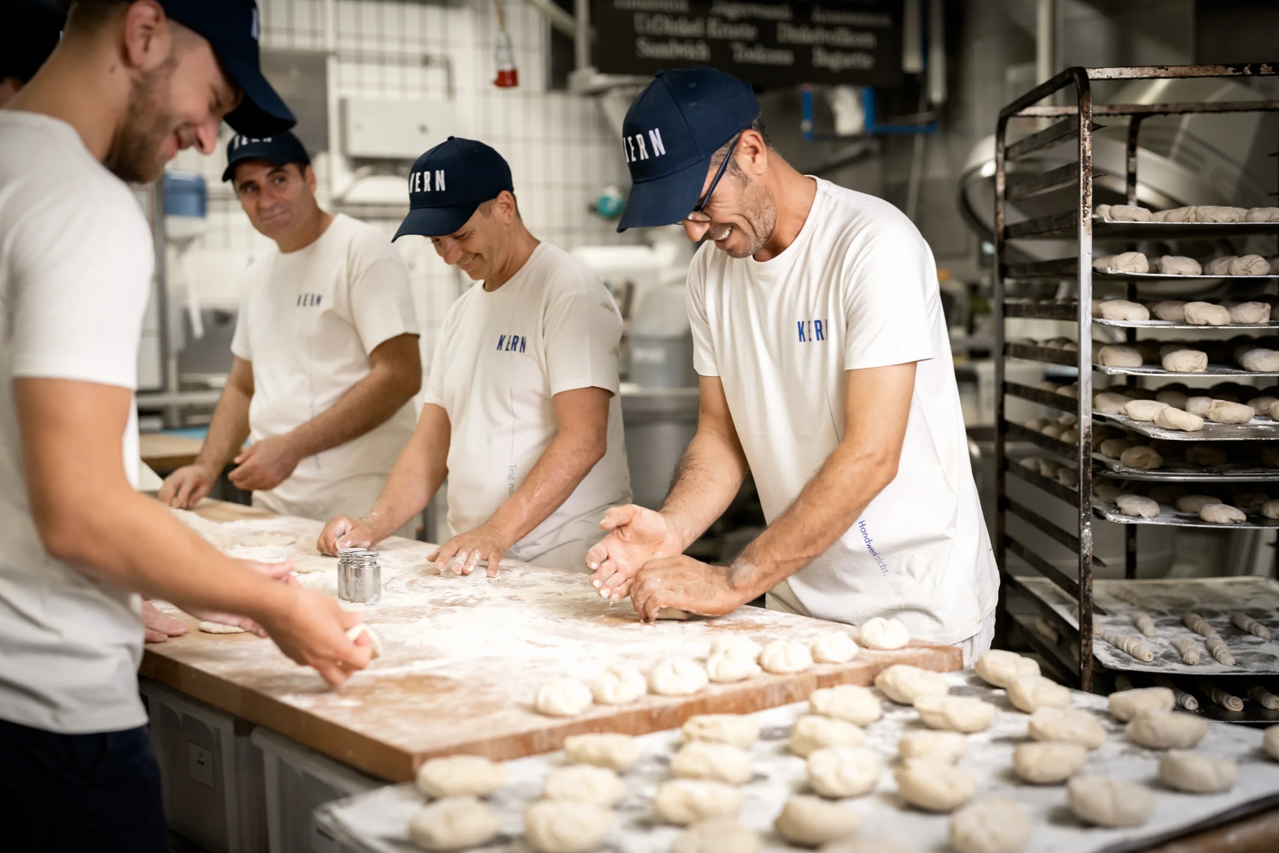 Vier Männer backen Teiglinge in einer Bäckerei, tragen weiße T-Shirts und blaue Caps, von denen eines 'KERN' steht. Auf dem Arbeitstisch liegen Teigstücke, und im Hintergrund sind Backwaren auf Metallregalen.
