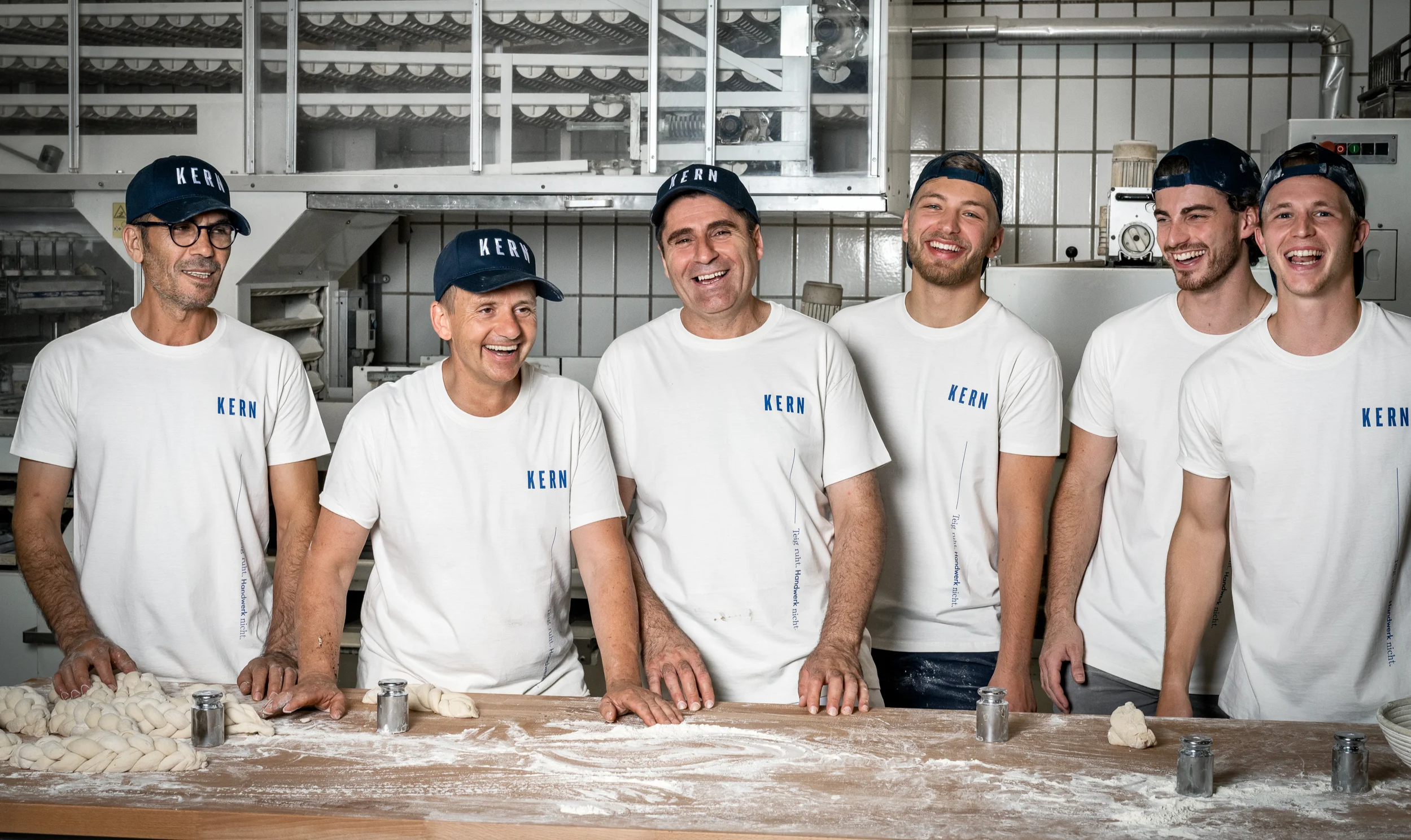 Sieben Männer in weißen T-Shirts mit dem Aufdruck 'KERN' und dunklen Caps lachen und arbeiten in einer Bäckerei. Der Tisch ist mit Mehl und Teigflecken bedeckt, auf dem Teiglinge liegen.