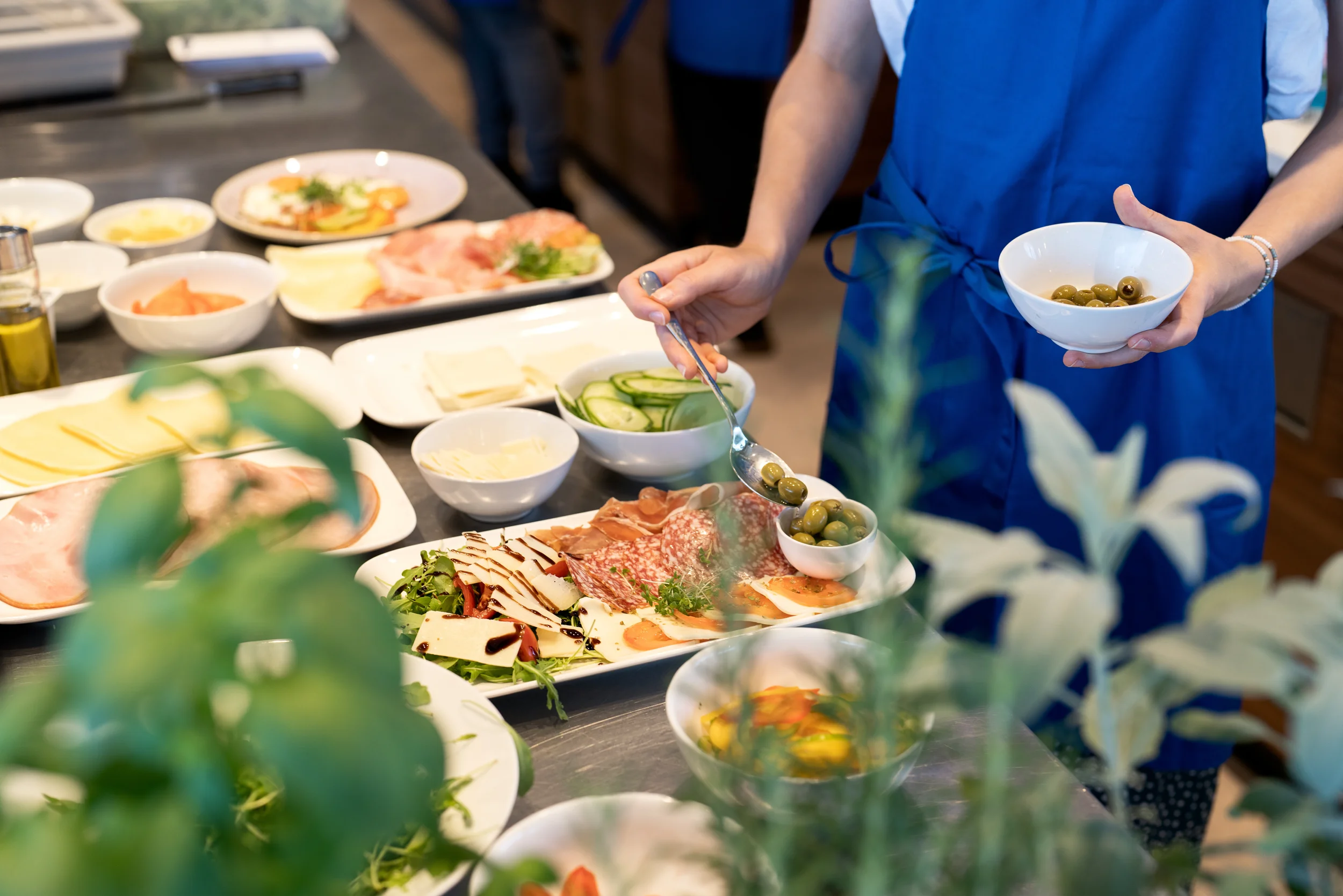 Person, die eine Schüssel mit grünen Oliven hält und Oliven auf eine andere Schüssel mit einem Löffel legt, umgeben von verschiedenen Speisen auf einem Buffet.