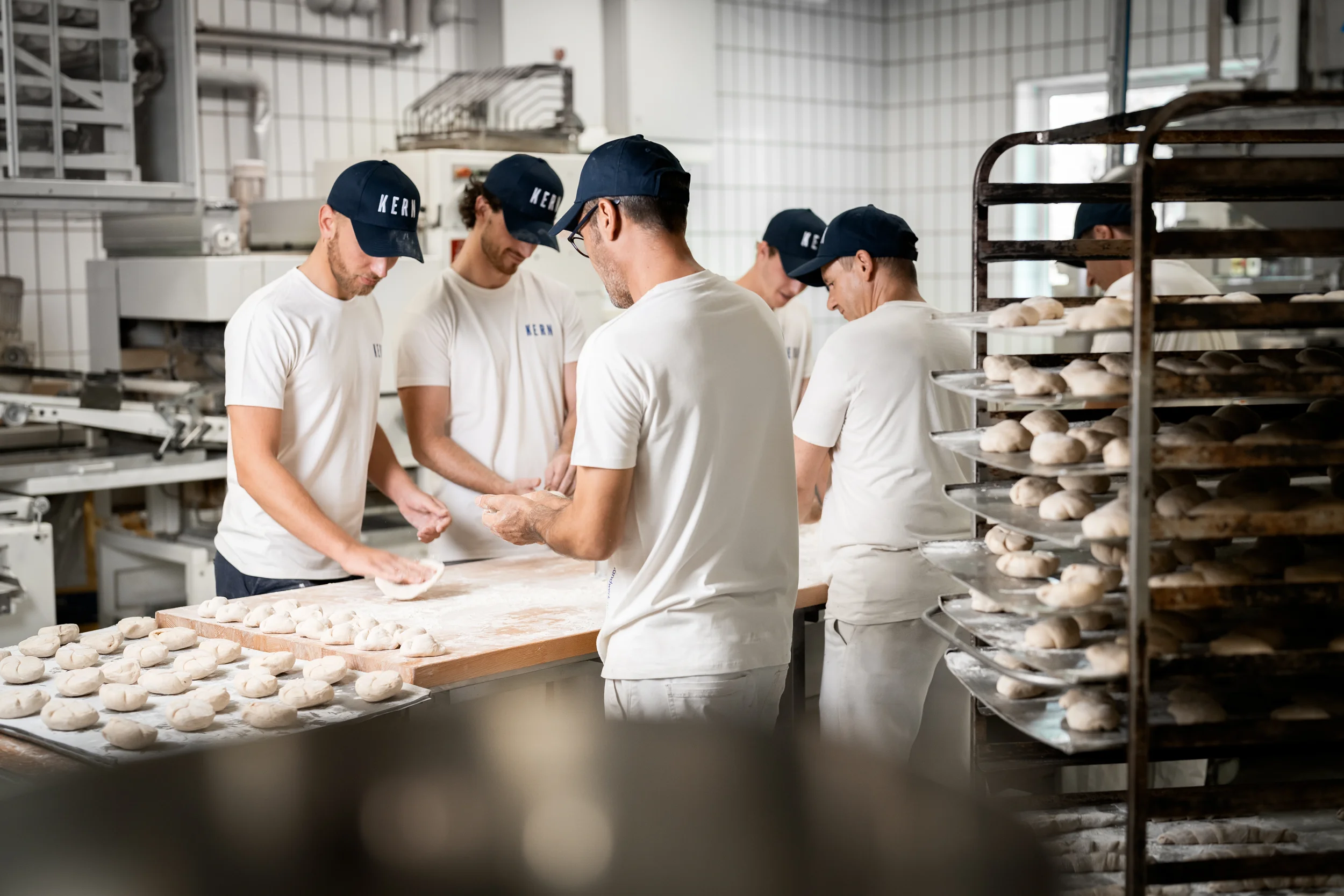 Menschen in einer Bäckerei beim Teig Kneten, mit mehreren aufgespießtem Teig auf Rosten im Hintergrund.