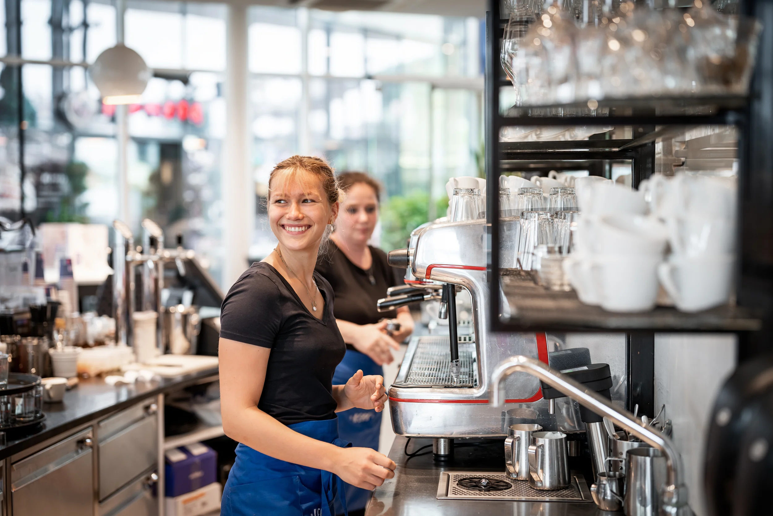 Lächelnde Barista in einem Café hinter einer Espressomaschine, mit einer weiteren Mitarbeiterin im Hintergrund.