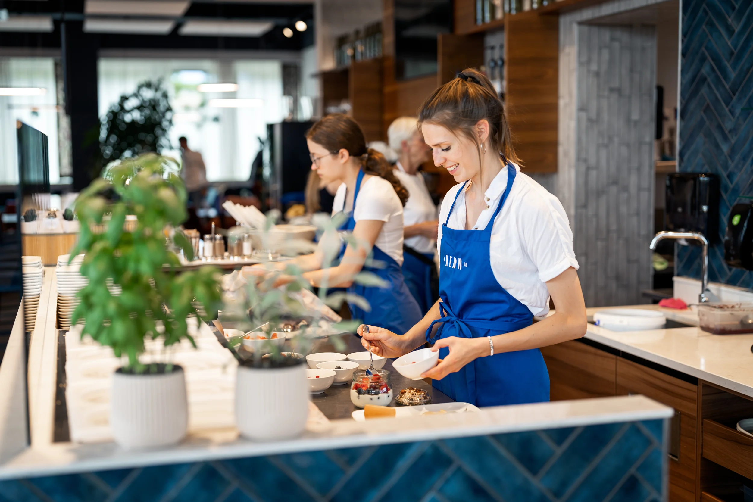 Mitarbeiter in blauen Schürzen bereiten in einer Küchenumgebung Desserts zu, während andere Mitarbeiter im Hintergrund arbeiten.