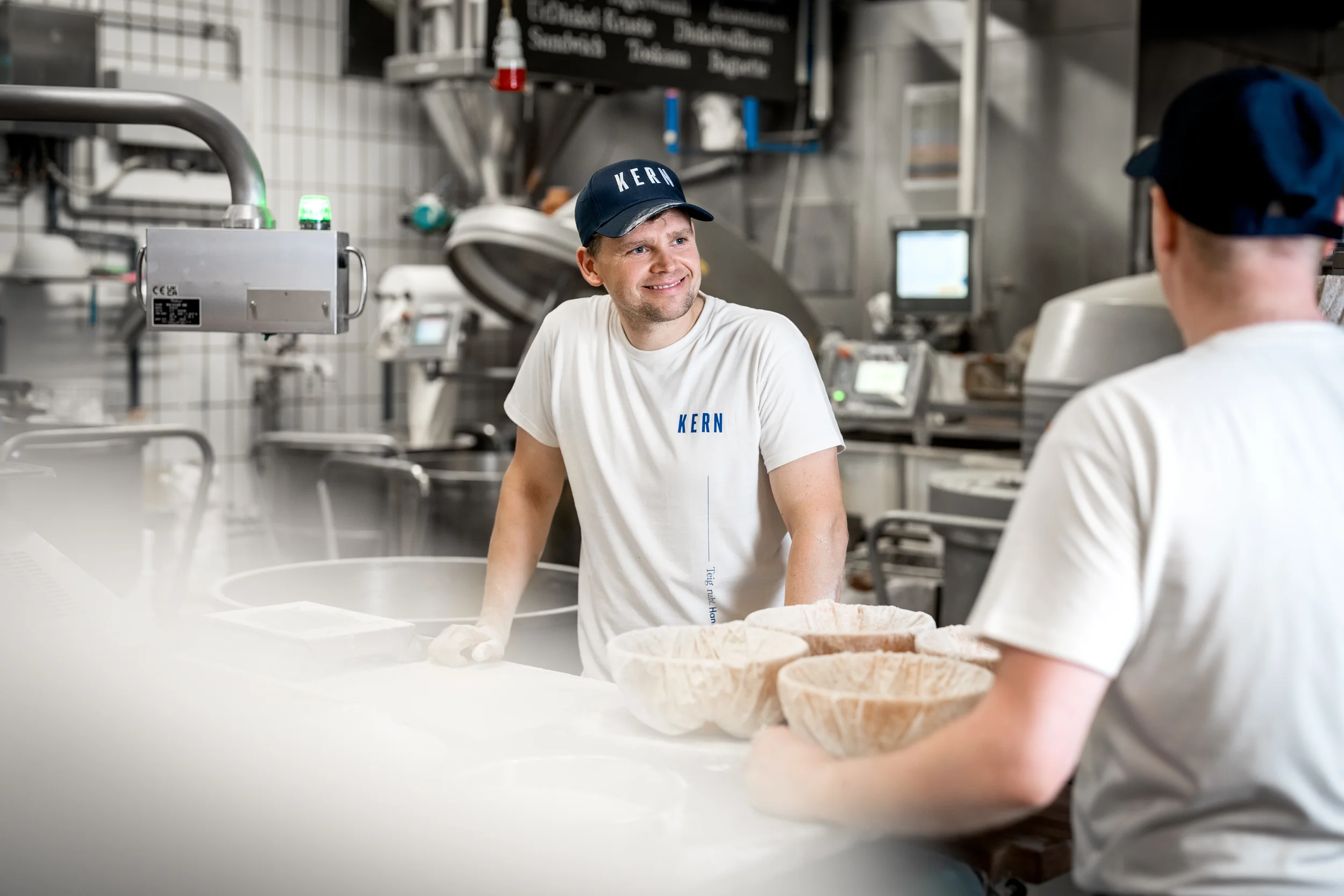 Zwei Männer in einer Industrieküche, einer lächelnd und der andere von hinten sichtbar, beide tragen weiße T-Shirts mit blauer Aufschrift "KERN" und blaue Kappen, die den Text ebenfalls zeigen, während sie mit großen Schüsseln arbeiten.