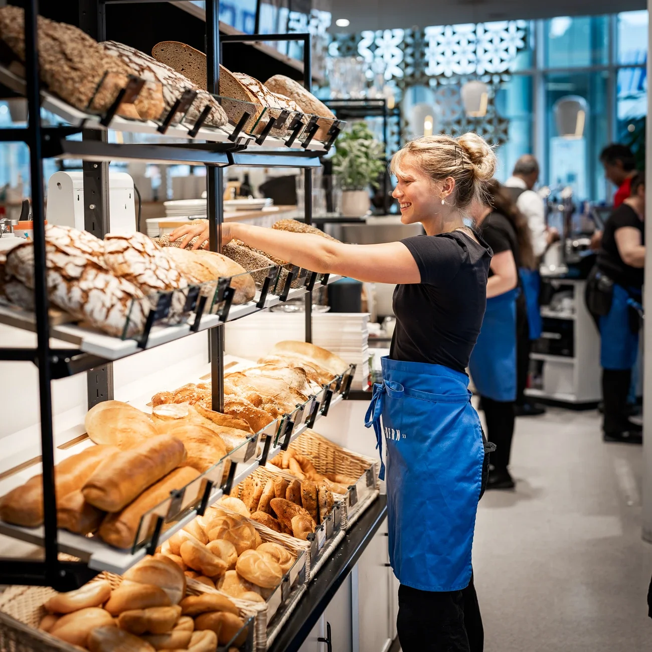 Eine Verkäuferin nimmt frisches Gebäck aus einem Regal in einer Bäckerei. Es gibt verschiedene Brotsorten und Brötchen, die auf mehreren Ebenen angerichtet sind.