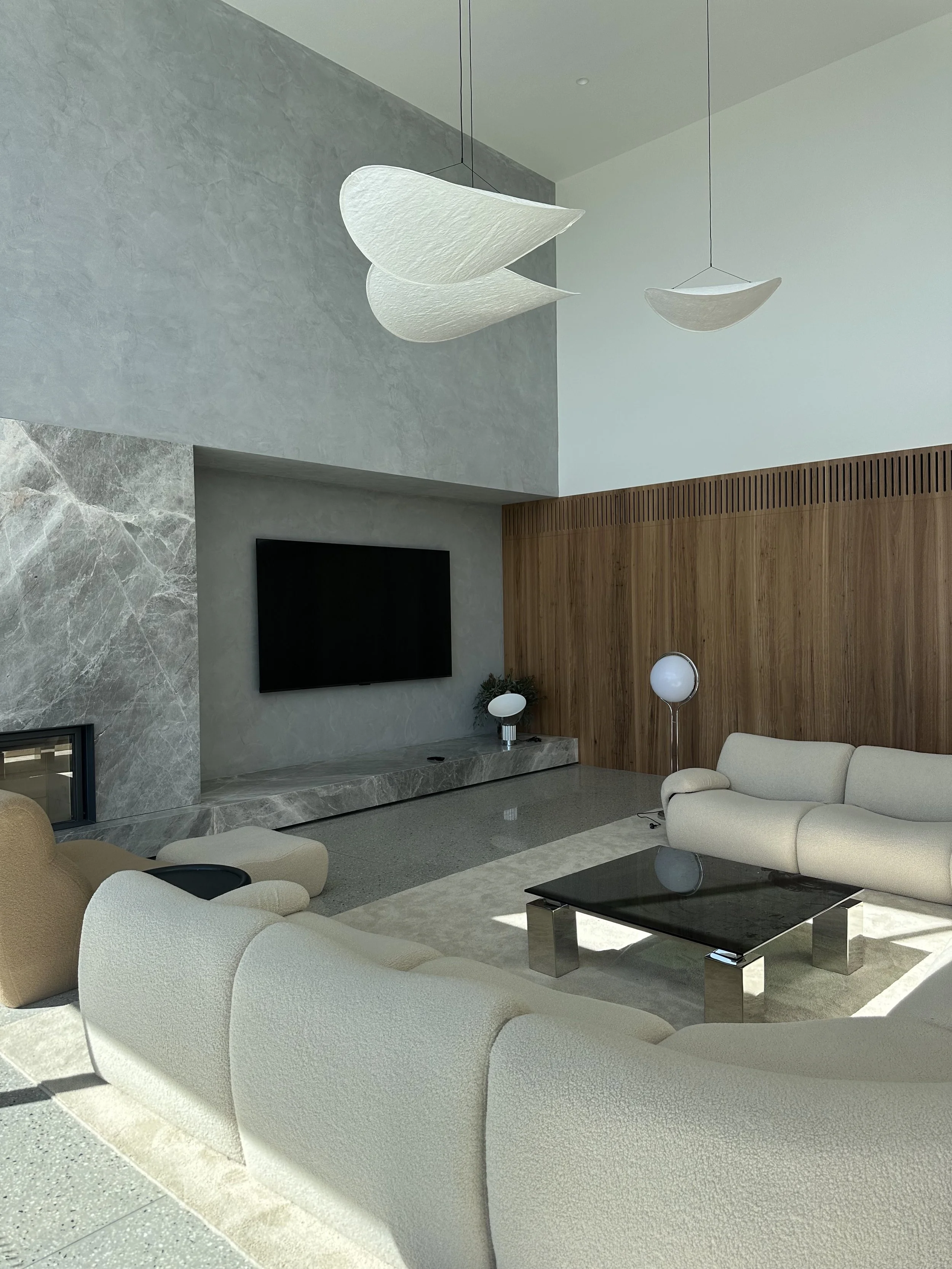 Modern living room with cream sofas, a black coffee table, a wall-mounted TV, decorative lamps, and a wooden-paneled wall in the background.