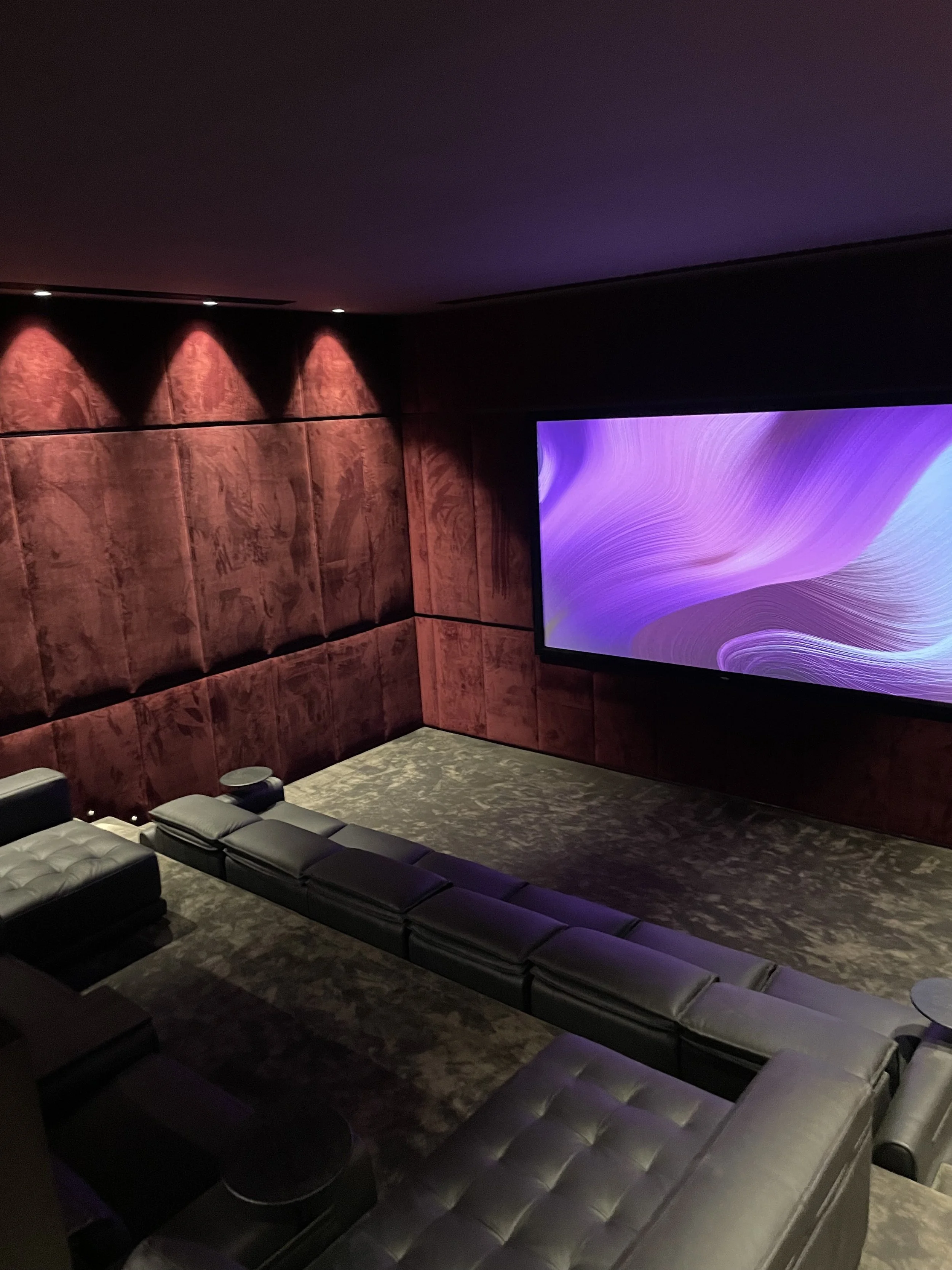 Luxury home theater room with plush black sofas arranged in a U-shape, a large abstract purple and blue screen on the wall, dark wood-paneled walls, and dim lighting.
