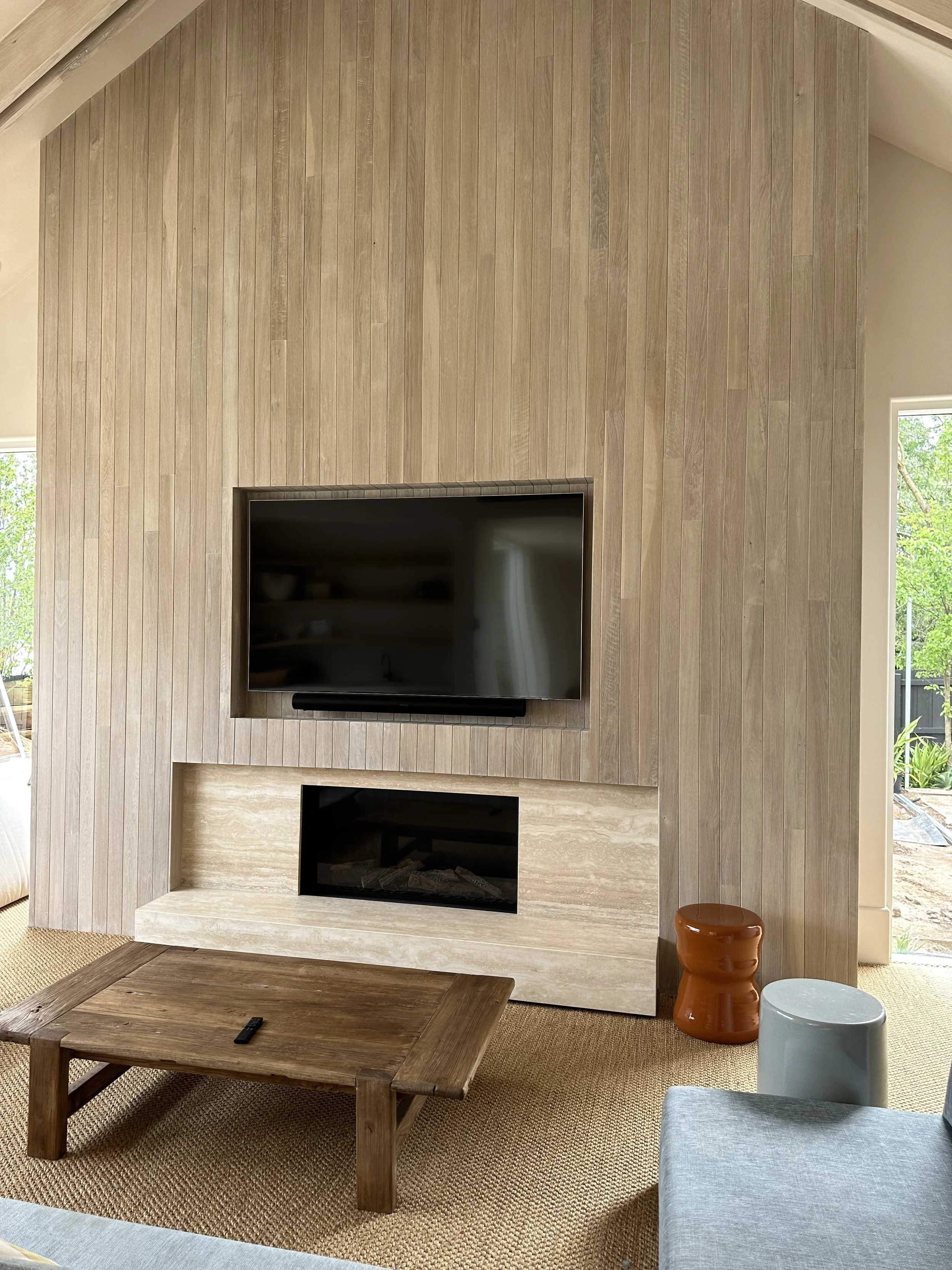 Living room with wooden accent wall, wall-mounted flat-screen TV, fireplace, low wooden coffee table, orange and gray ceramic stools, and part of a blue sofa.