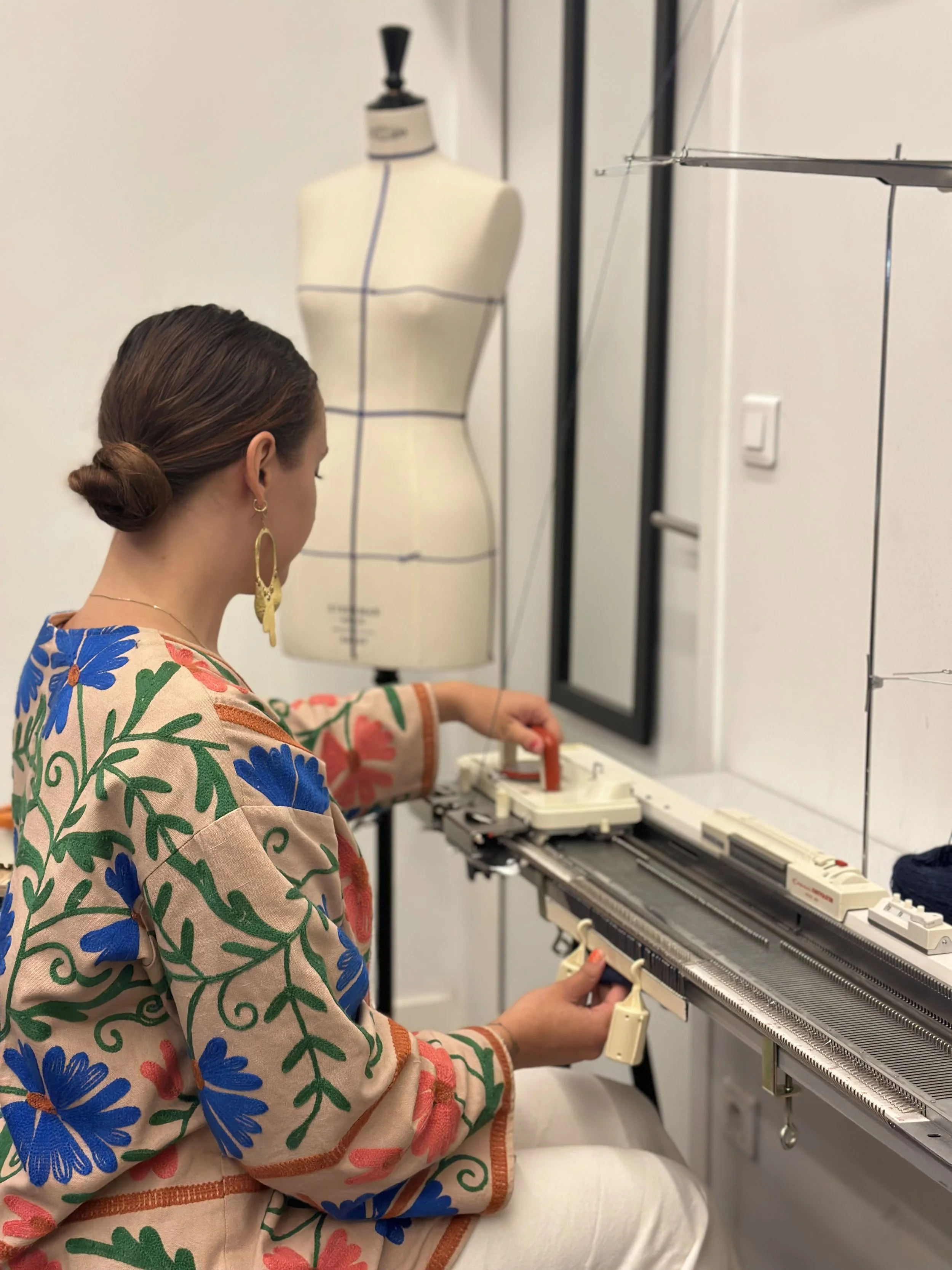 Une femme en vêtement coloré travaille avec un piano a tricoter. Elle créer de nouveaux tricot  avec un mannequin en arrière-plan.