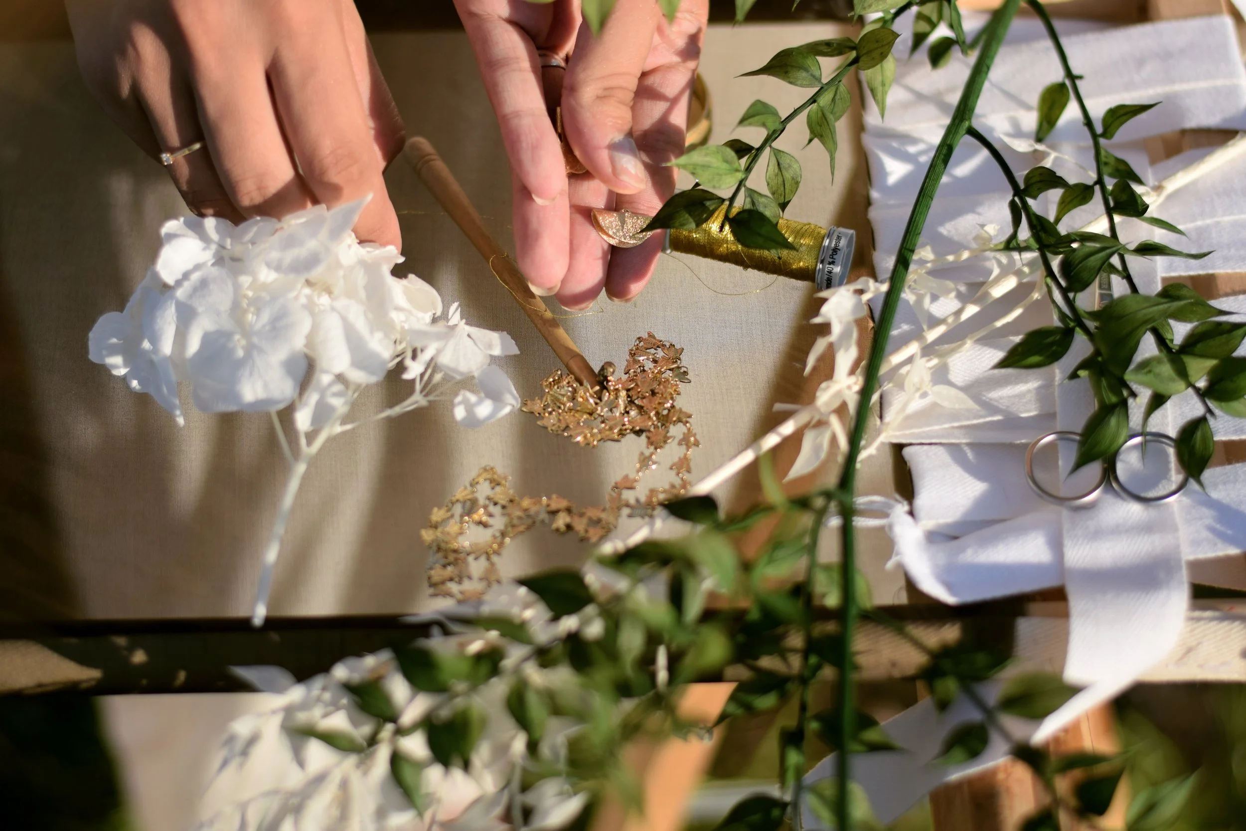Mains manipulant une branche de plante verte avec des feuilles de différentes nuances de vert et blanc, entourée de diverses décorations dorées et blanches sur un métier a broder. On voit aussi des fleurs blanches stabilisé.