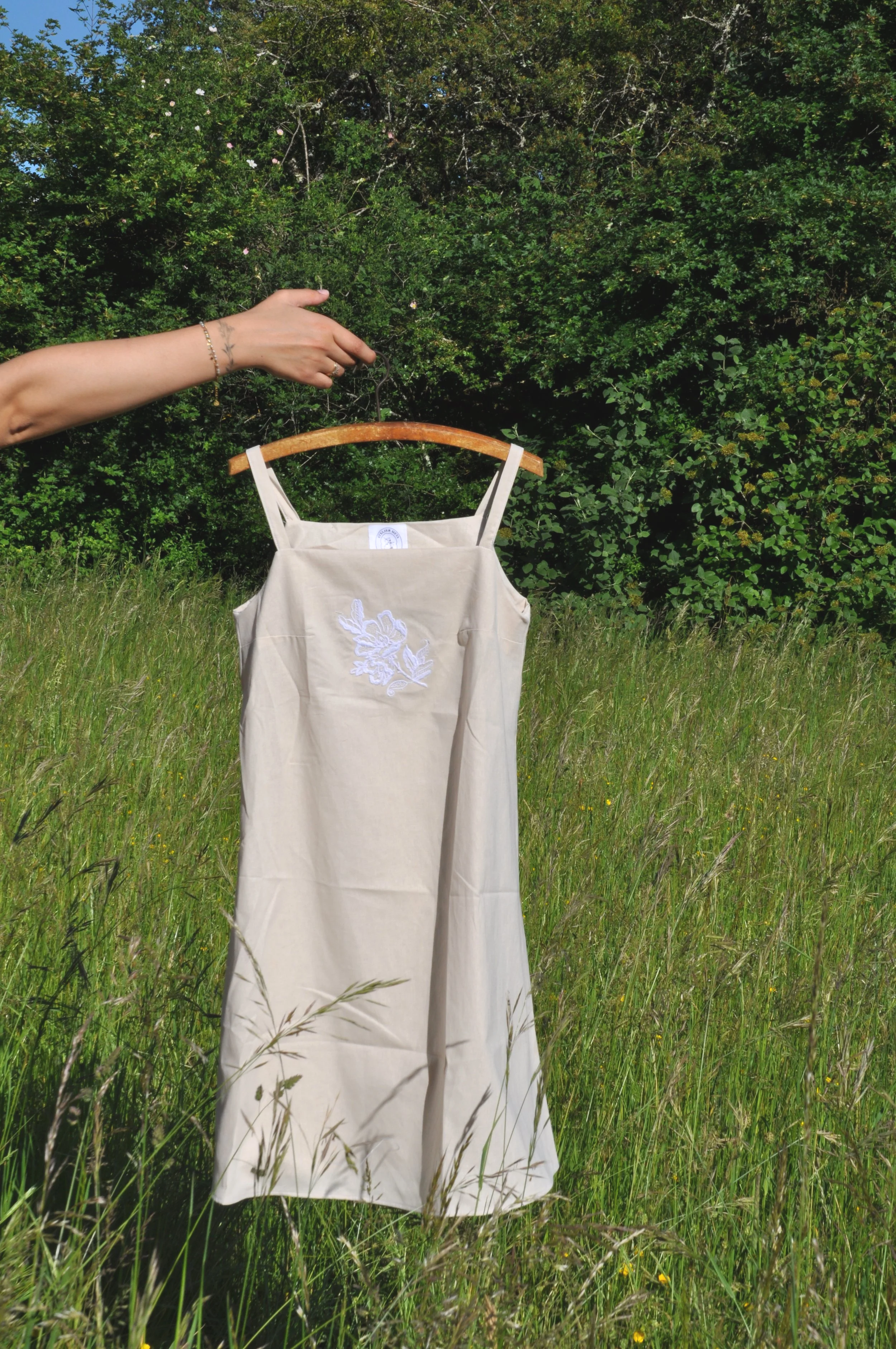 Une main tient un cintre avec une robe beige en plein air, forêt en arrière-plan.