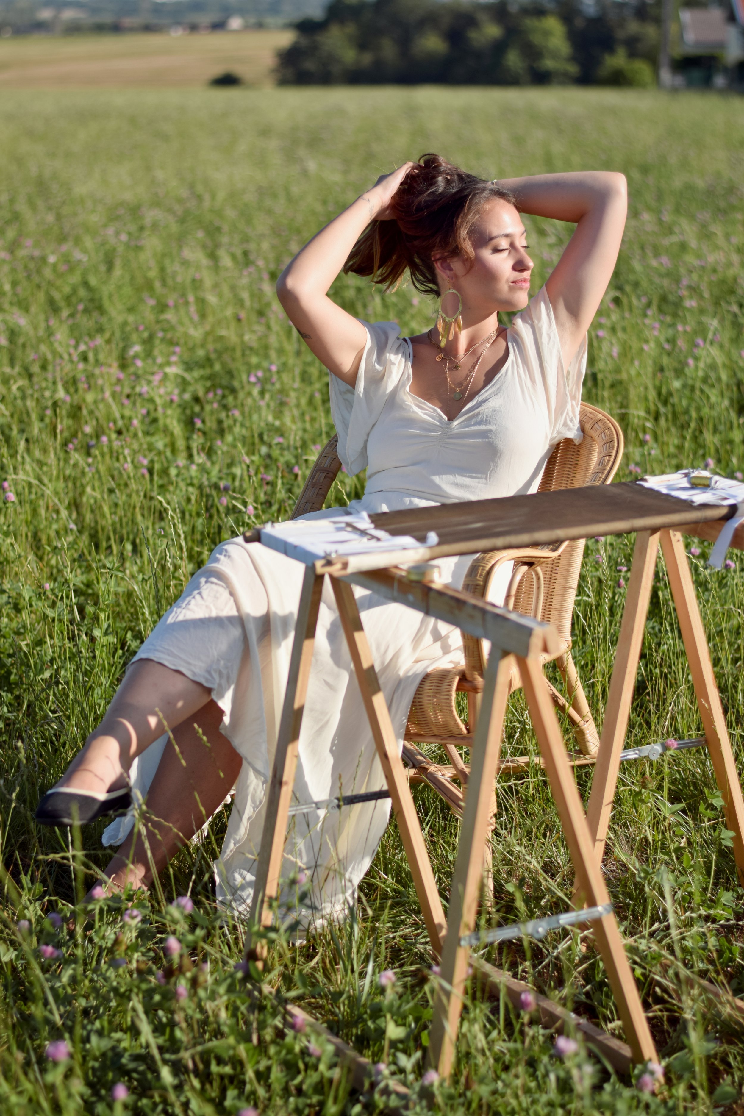 Femme assise dans un champ de fleurs, profitant du soleil avec les yeux fermés, mains dans les cheveux, portant un vêtement blanc et de grands accessoires.