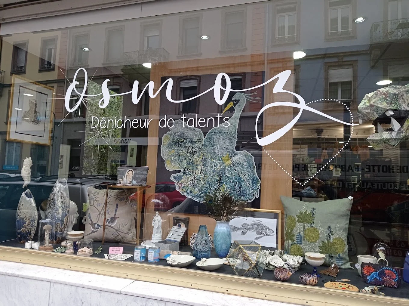 Vitrine d'une boutique intitulée 'Osmose, dénicheur de talents', affichant divers objets décoratifs, peintures, vases, coussins, et une illustration d'un héron avec un cœur en pointillés.