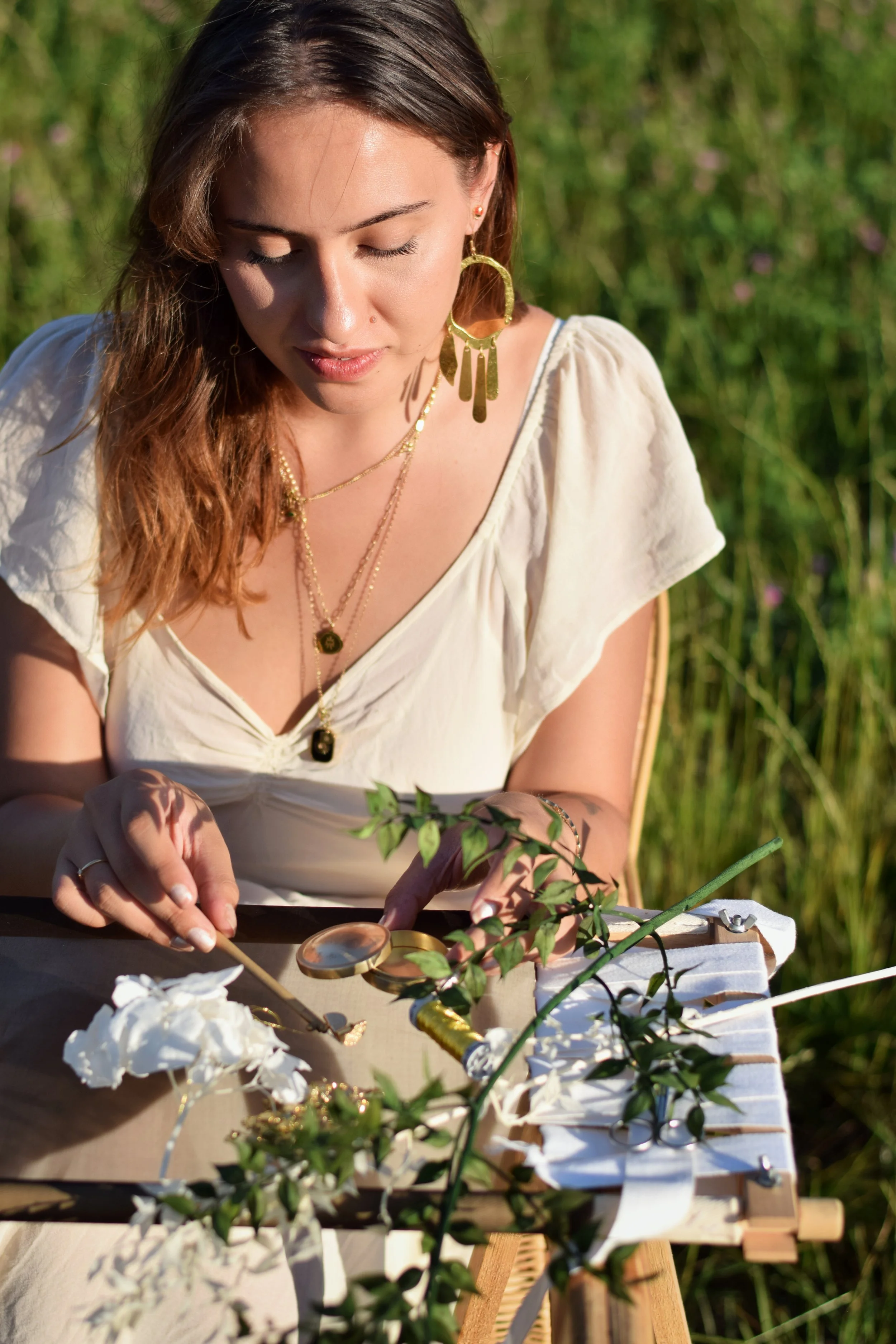 Une femme portant des bijoux dorés, un chemisier blanc, examine des plantes ou des feuilles sur son métier a broder, dans un environnement extérieur verdoyant.