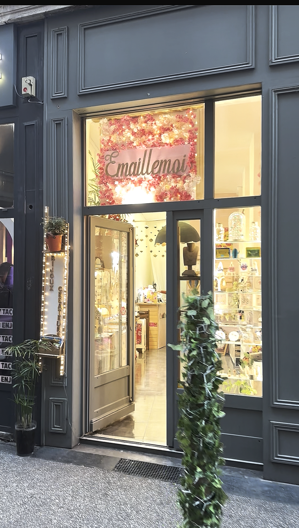 Vitrine d'une boutique avec un panneau marquée 'Enallemoi', décorée de fleurs roses et blanches, avec une porte en verre ouverte, intérieur éclairé avec des étagères contenant divers objets.