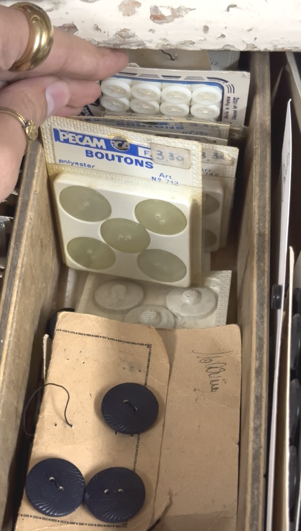 Un tiroir en bois contenant des boutons de différentes tailles, couleurs et matériaux, parmi lesquels des boutons noirs et blancs, dans des emballages en carton et en plastique.