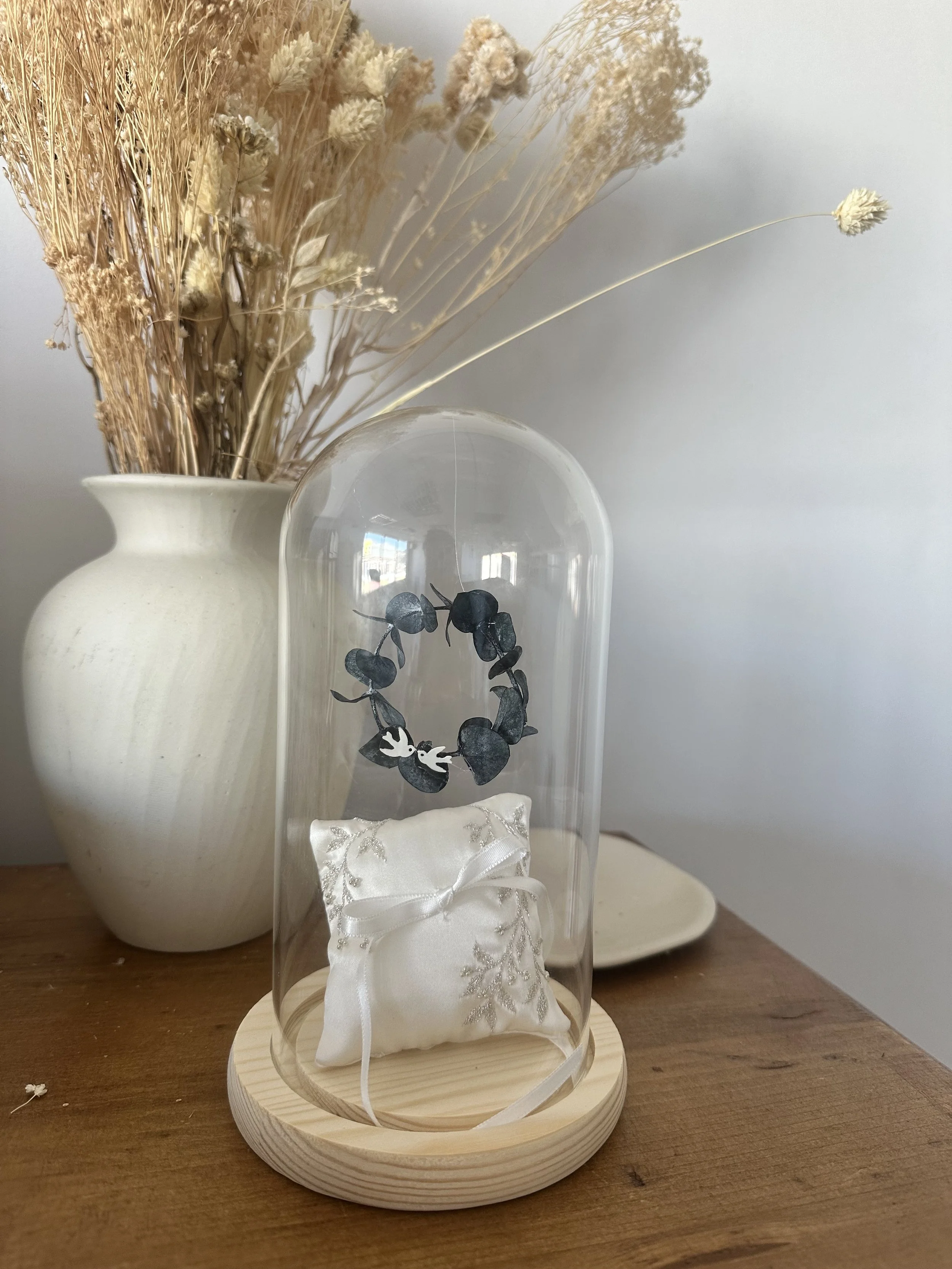 Un vase blanc contenant des fleurs séchées de couleur crème à l'arrière-plan, et une boîte en tissu blanc avec un ruban en satin et une broderie dorée à l'avant, posées sur une table en bois.