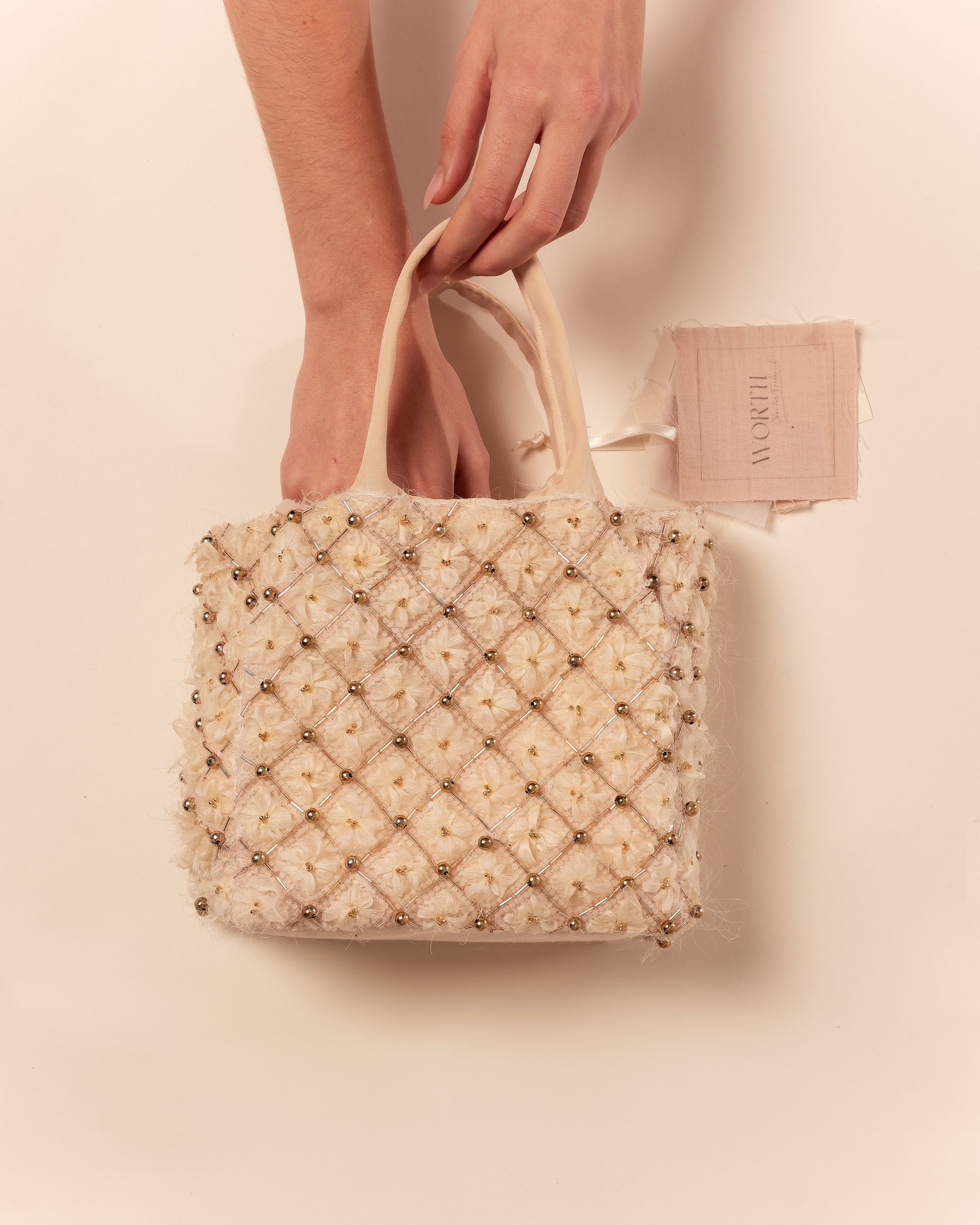 Un sac à main orné de perles et de fleurs en tissu, posé sur une surface claire avec une étiquette en tissu rose à côté.
