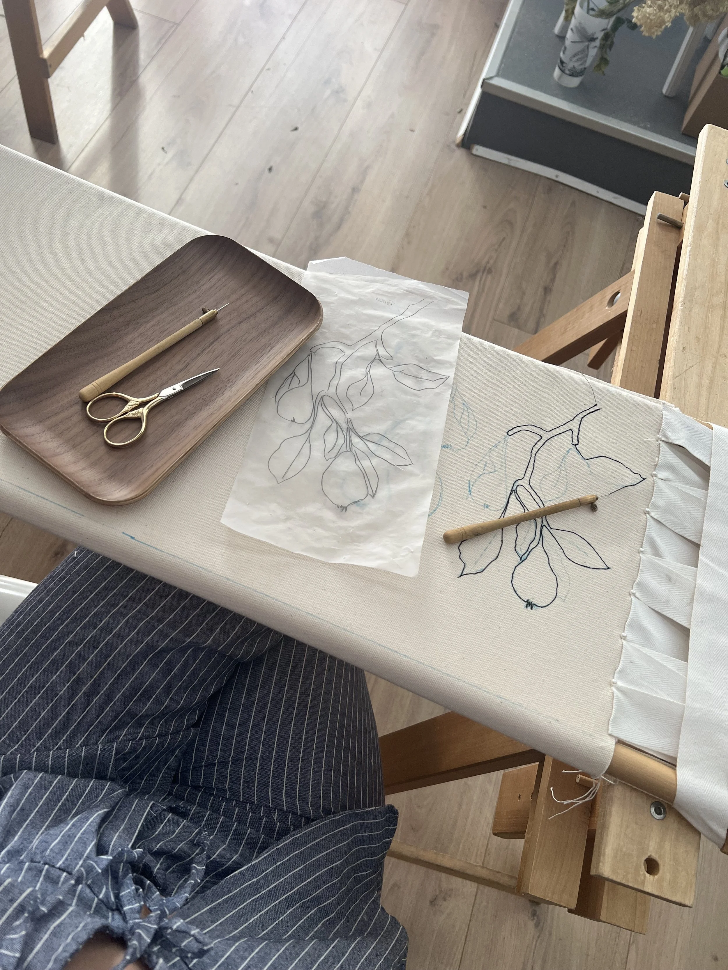 Métier a broder avec deux croquis de branches et feuilles. Outils de broderie, ciseaux. Personne portant un pantalon à rayures assise devant le métier a broder.