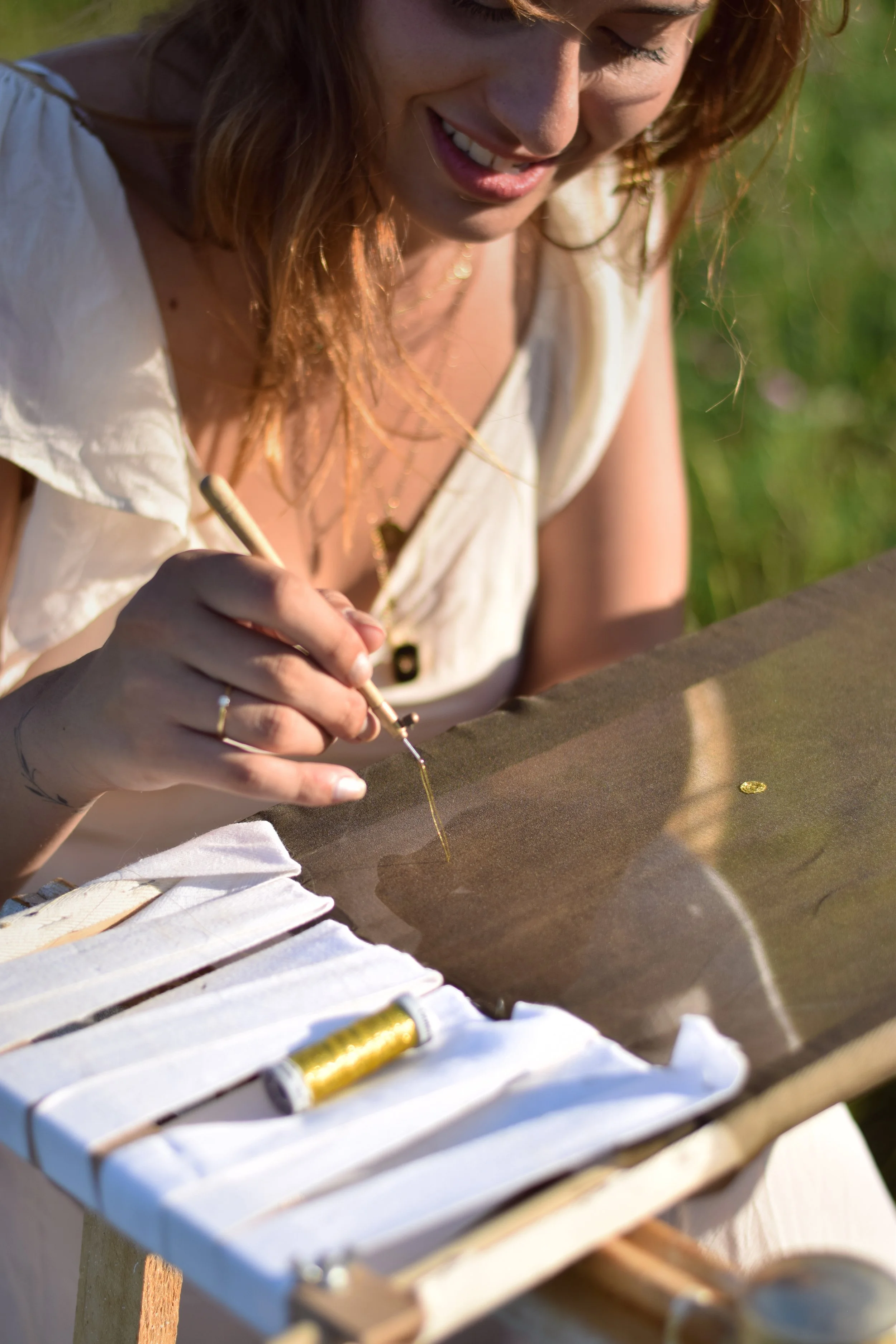 Une femme est assise devant sont métier a broder en extérieur, concentrée sur la création de broderie avec un outil fin (crochet de Lunéville). Sur la table, il y a des bobines de fil doré et des fournitures de bijouterie.
