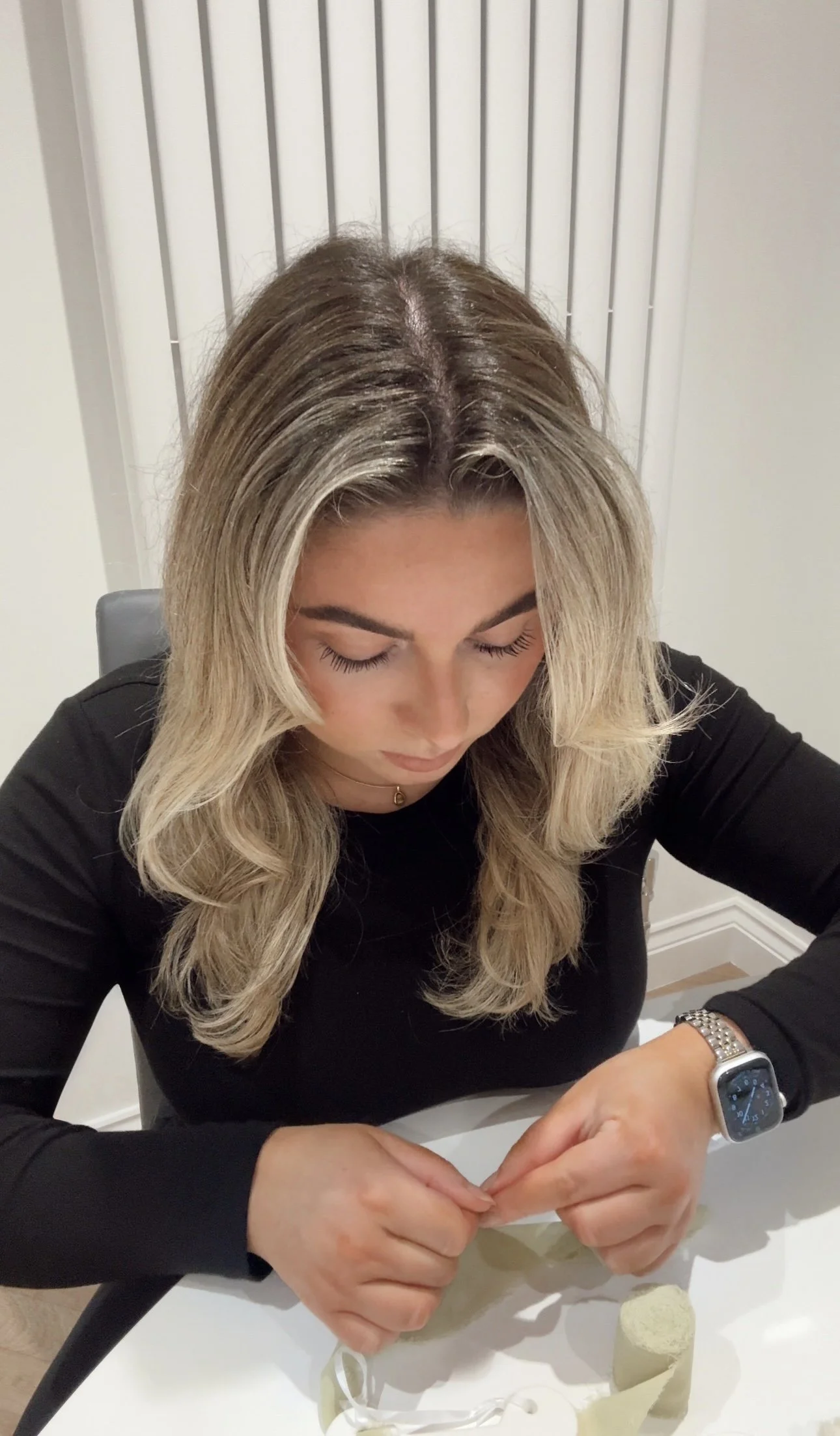 A woman with blonde hair, wearing a black top and a watch, is sitting at a table with her head bowed down, engaged in calligraphy involving ribbon, brush pens and ceramic baubles.