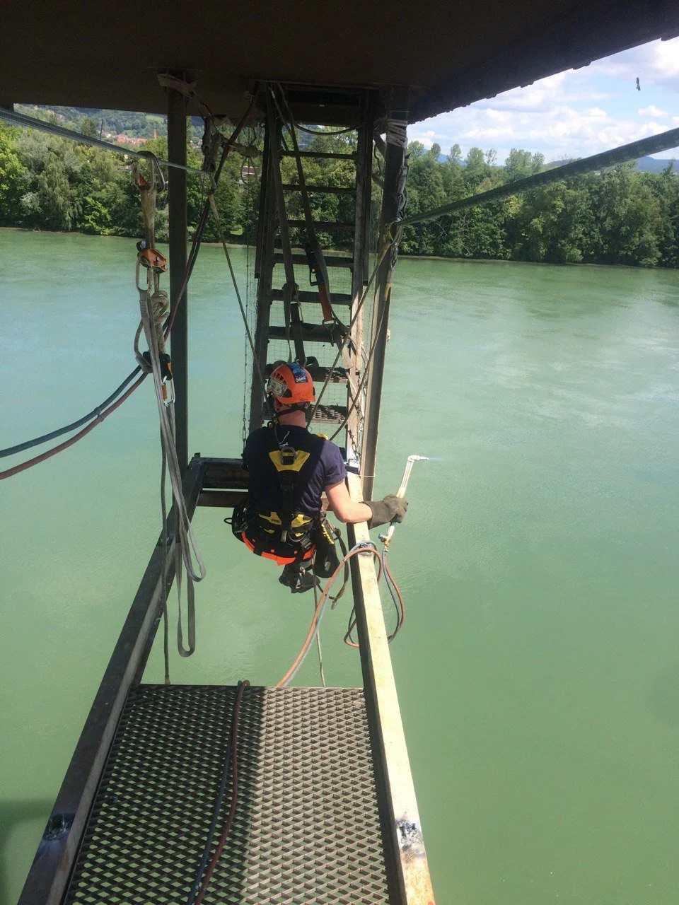 Ein Kletterer hängt an einem Seil an einer Metallleiter über Wasser mit grüner Uferlandschaft im Hintergrund.