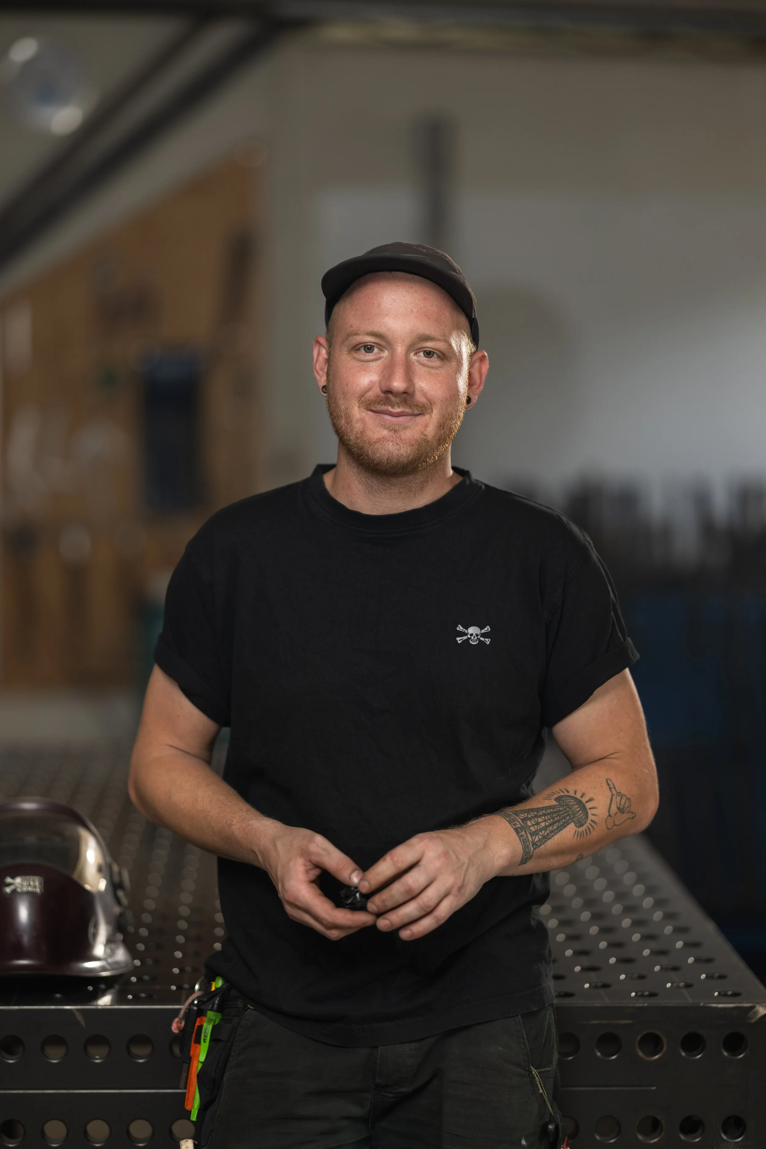 Ein junger Mann mit rotem Bart und Tattoos, trägt ein schwarzes T-Shirt und eine schwarze Mütze, steht vor einer Werkbank in einer Werkstatt.