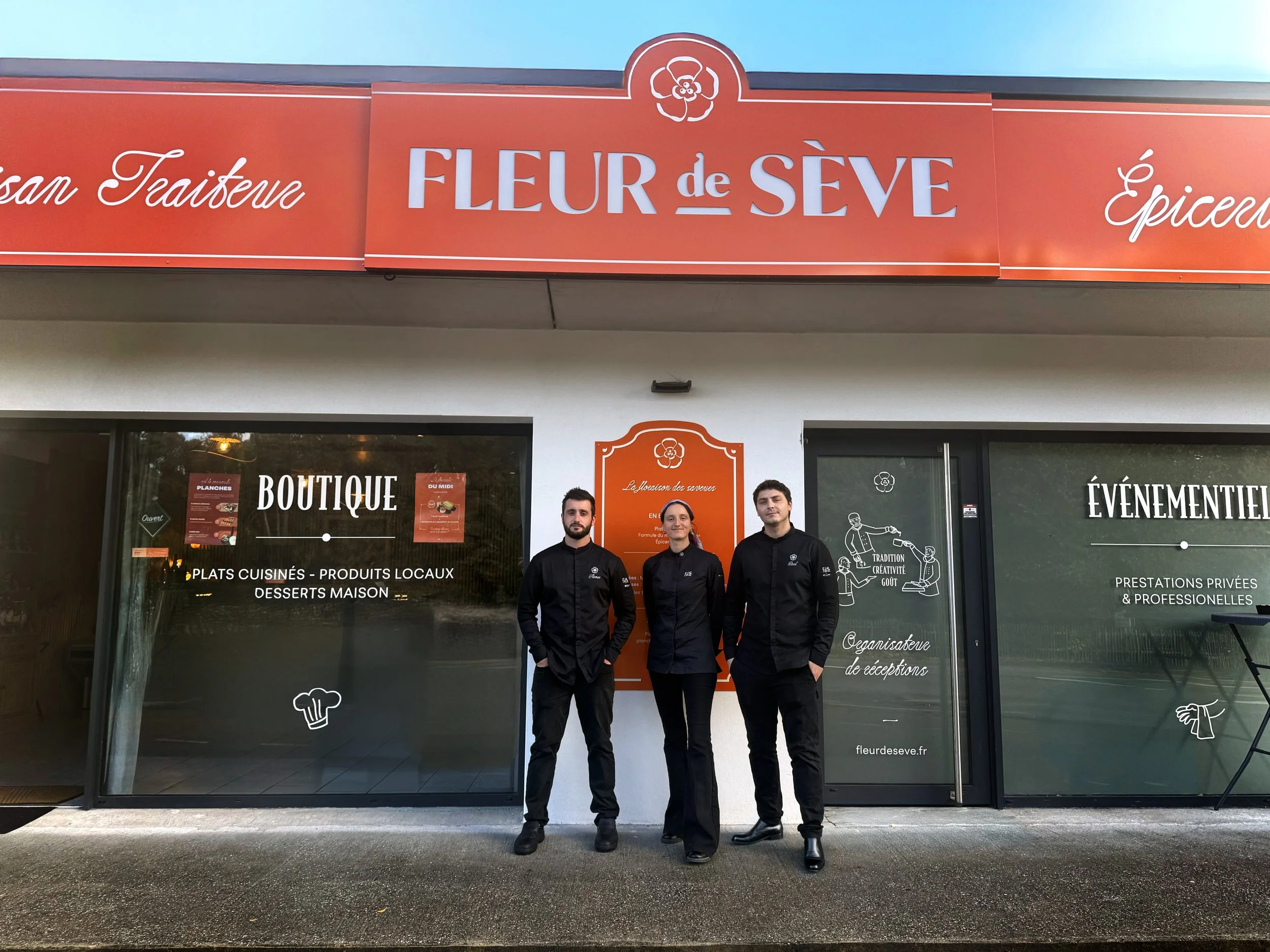 L'équipe de Fleur de sève en tenue noire à savoir les trois gérants posant devant la façade de la boutique Fleur de Sève, avec des panneaux en vitrine affichant des menus et des informations.»