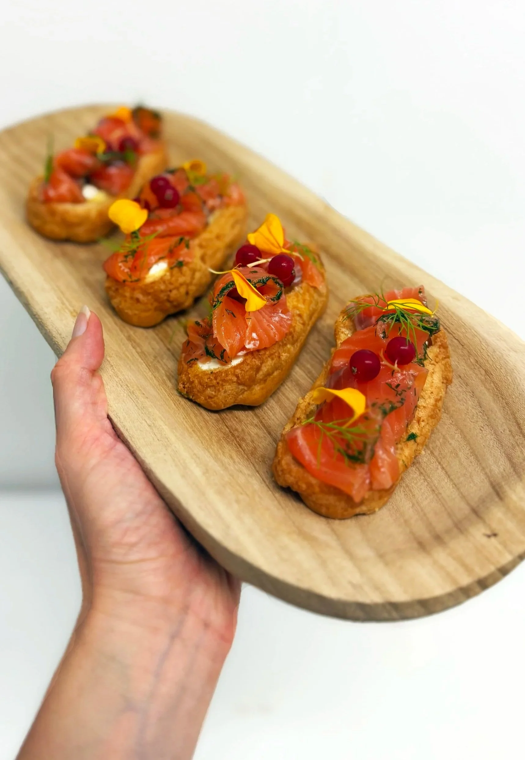 Eclairs salé au saumon fumé, groseille, pousse et fleurs comestibles, présentés sur une planche en bois.