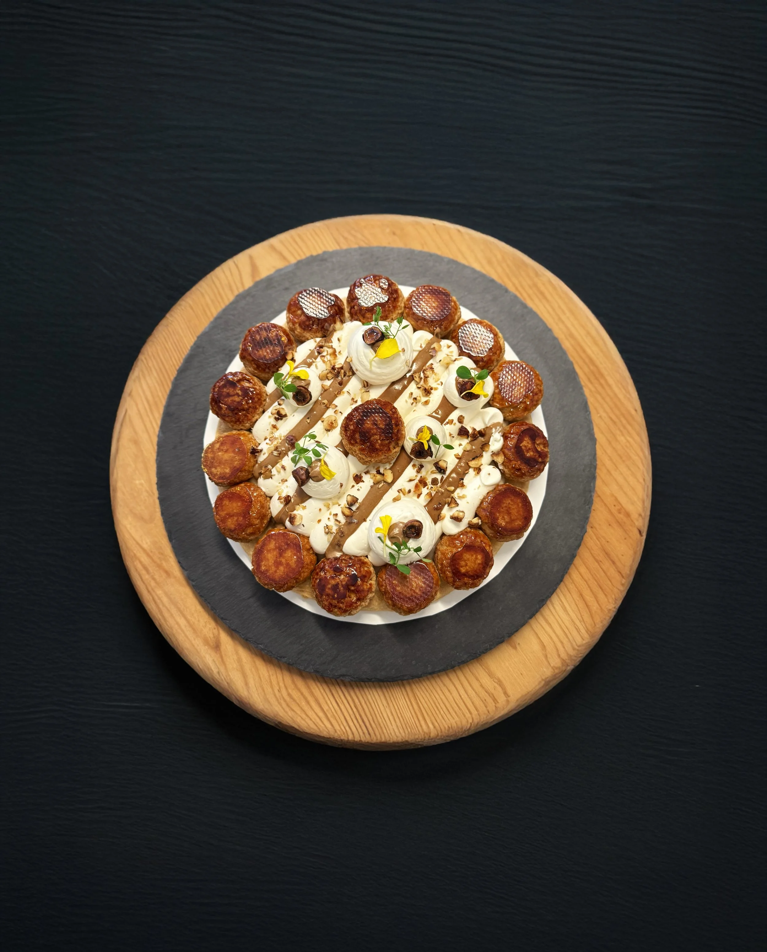 Saint-Honoré (gâteau composé de petits choux) élégamment décoré de fleurs et présenté sur une assiette en céramique noire sur un plateau en bois.