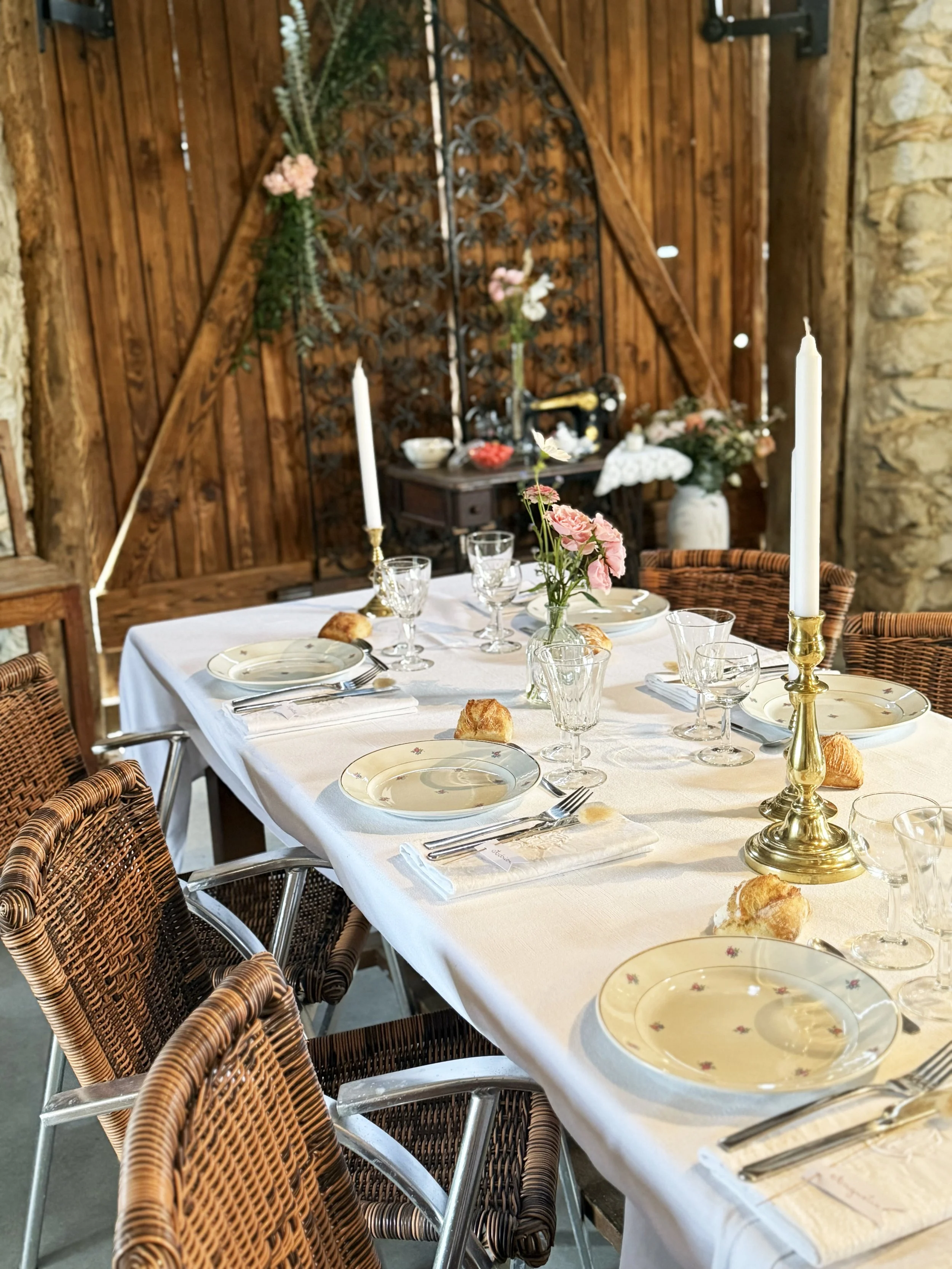 Table de repas élégamment dressée avec une nappe blanche, des assiettes, des verres à vin et des bougeoirs avec des bougies, décorée de fleurs roses, dans un cadre rustique avec un mur en pierre et une porte en bois.
