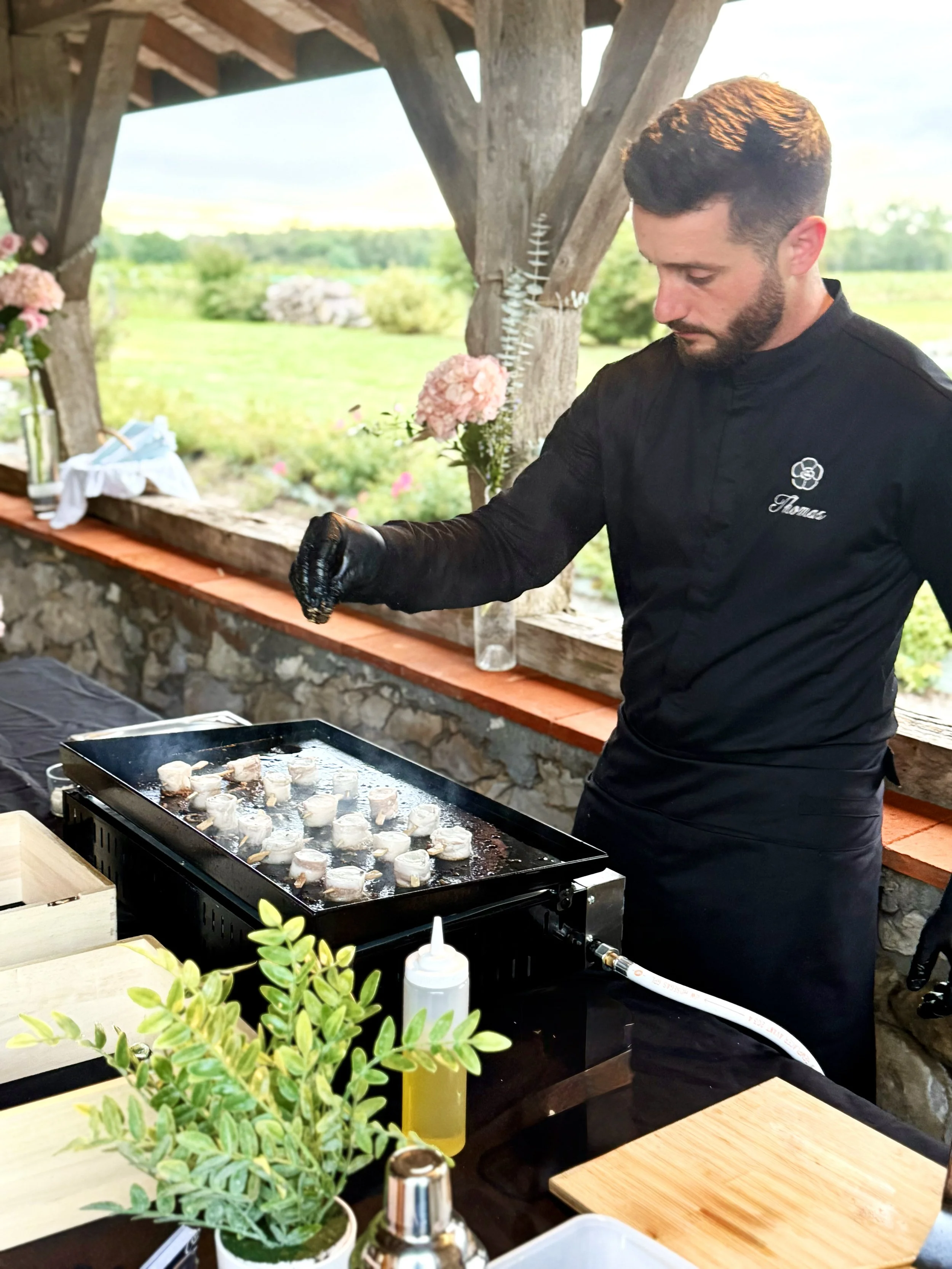 Un chef en extérieur prépare les brochettes de lotte au lard sur une plancha, entouré d'un décor champêtre avec des fleurs et un paysage verdoyant.