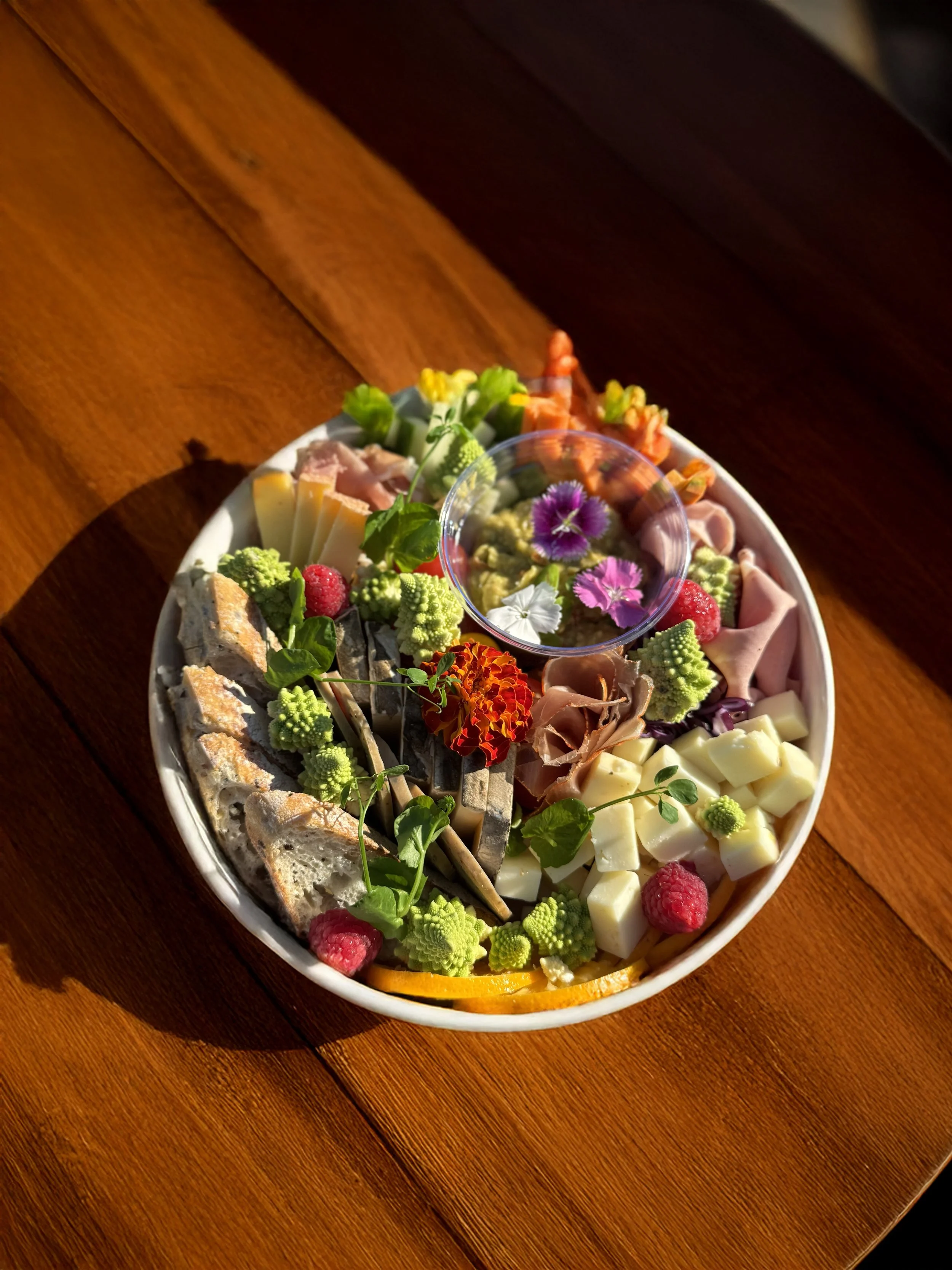 Box d'apéritive avec fromages, charcuterie, légumes croquants, toast, puis fruits et fleurs comestibles en décoration, présentée sur une table en bois, avec une lumière naturelle chaude.