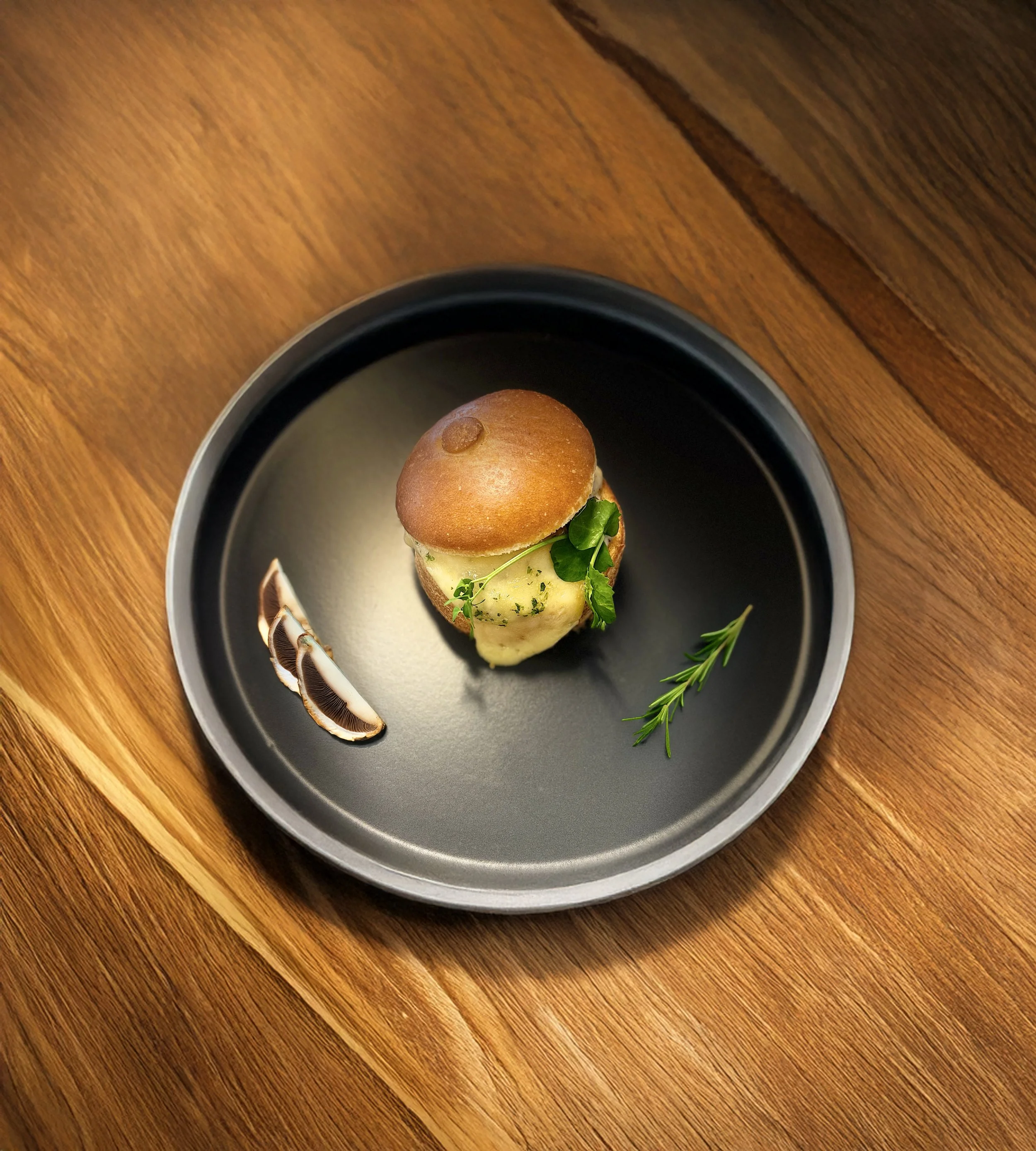 Brioché garni au poulet et champignon et gratiné au fromage à raclette. Il est décoré d'une pousse de petits pois avec une branche de romarin à côté, présenté dans une assiette noire sur une table en bois.