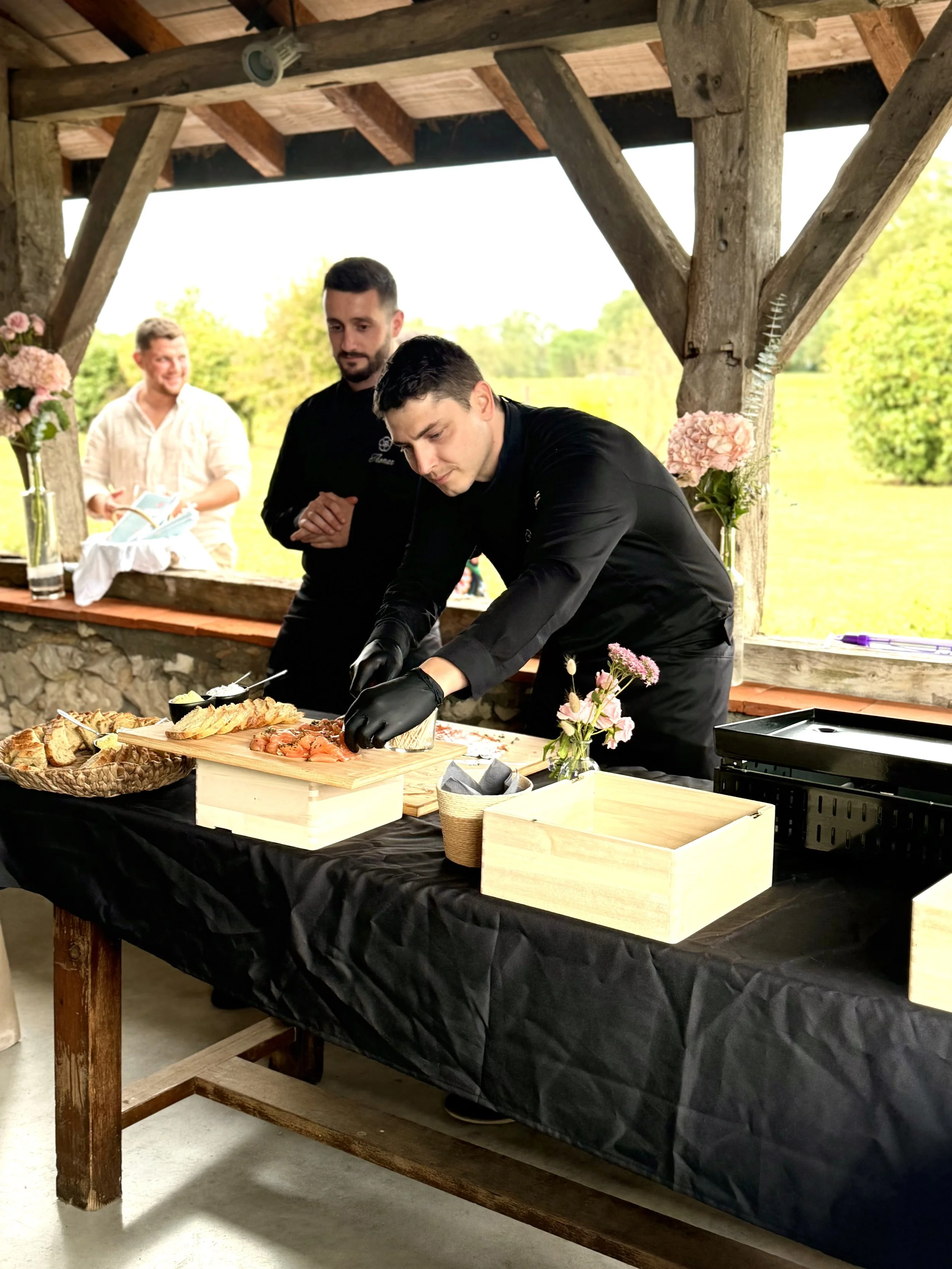 Un chef en noir prépare du saumon gravlax sur une table noire sous une structure en bois, avec deux autres personnes qui l'observent. La scène se déroule dans un espace en plein air avec un paysage verdoyant en arrière-plan.