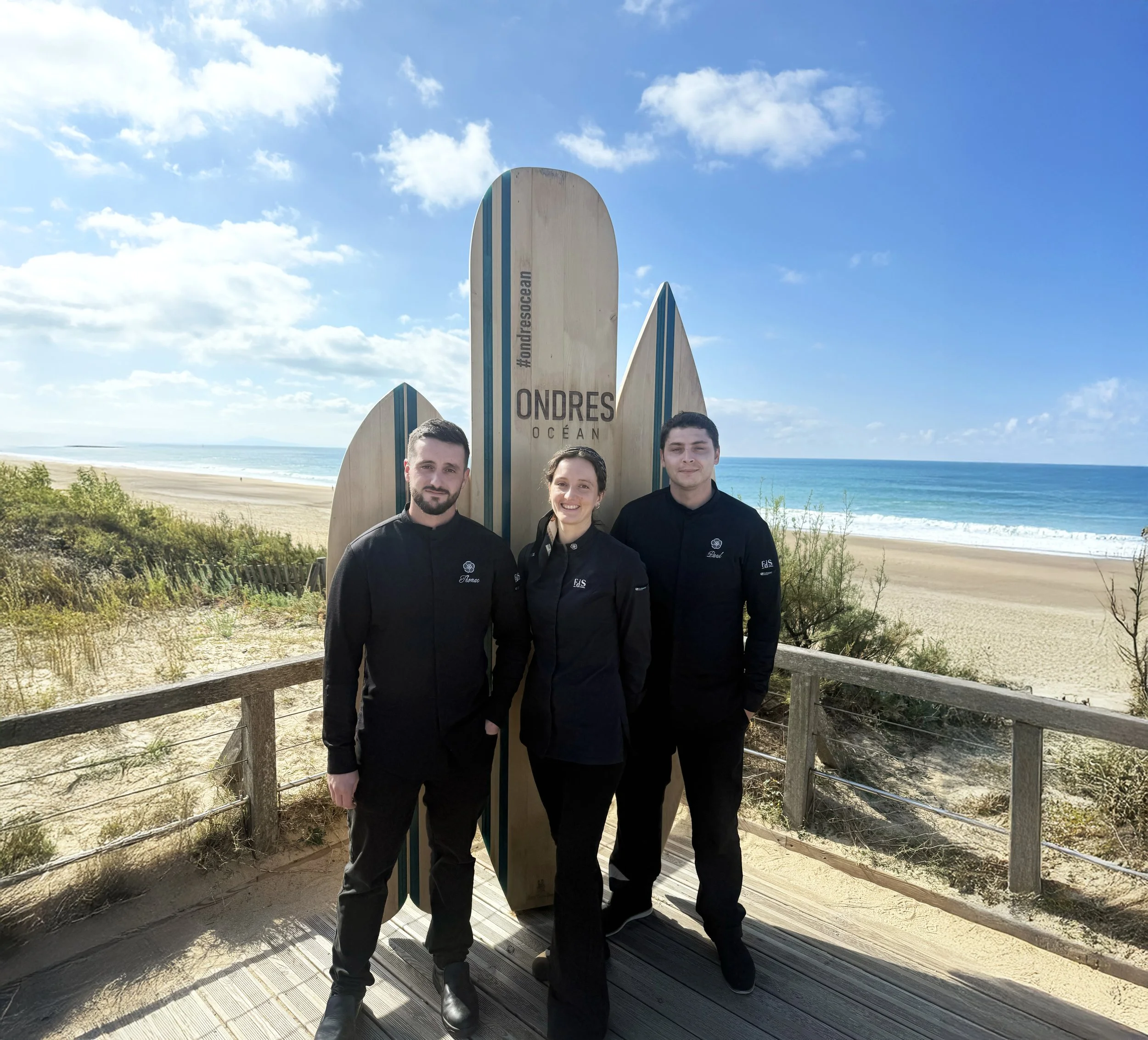 Les deux chefs et la cheffe de projets habillées en uniformes noirs posent devant la plage d'Ondres. Derrière eux, des planches de surf dont une indiquant 'Ondres Océan'.