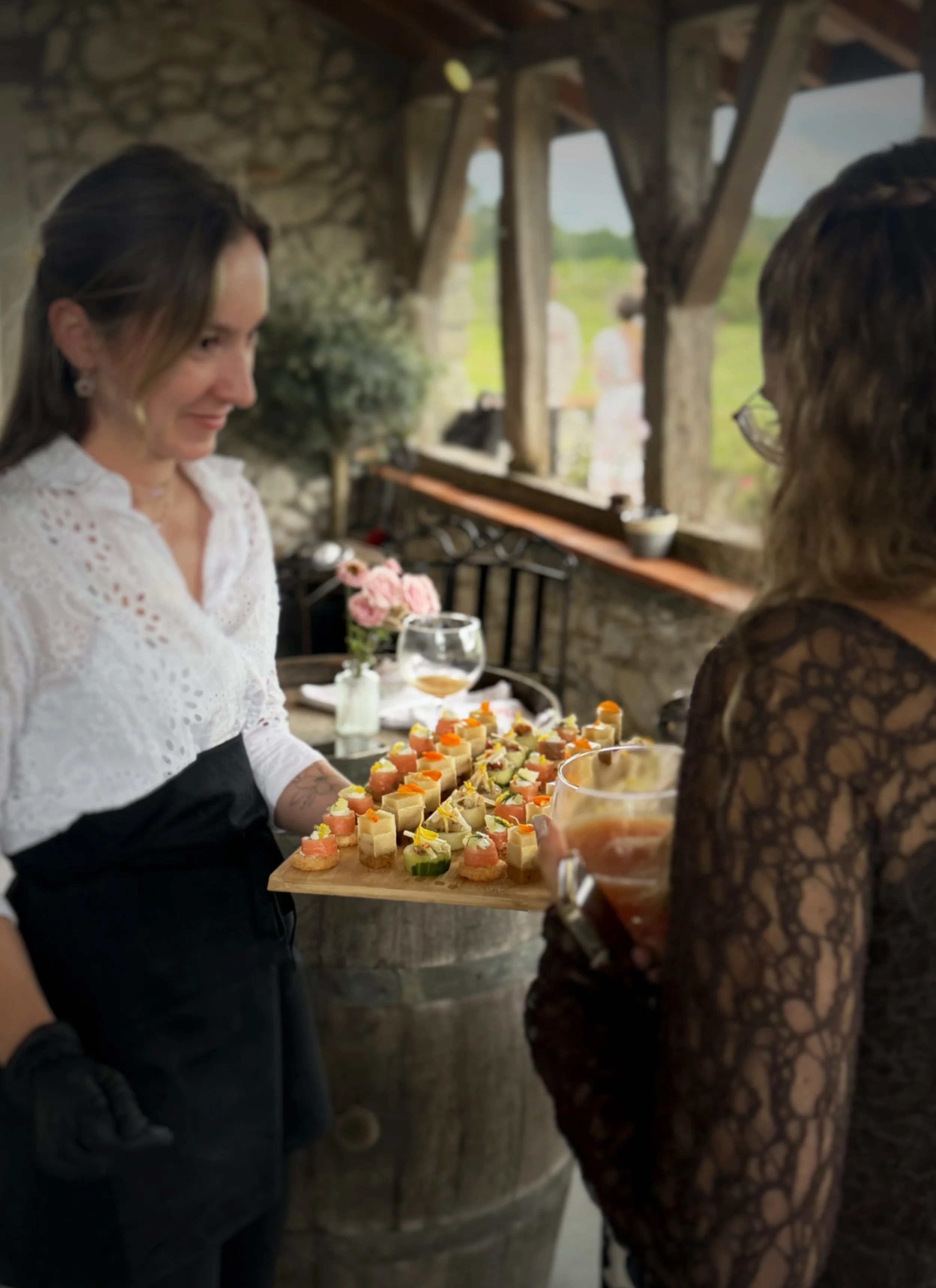 Deux femmes discutent lors d'une réception, une d'entre elles tient une planche en bois avec nos pièces cocktail froides. La scène se déroule dans une ambiance rustique avec un mur en pierre et une vue sur la nature en arrière-plan.