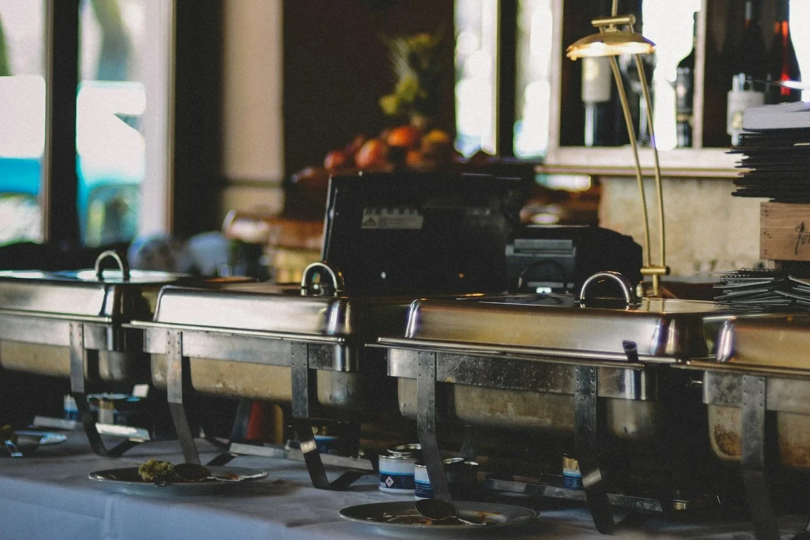 Buffet table setup with chafing dishes, a rice cooker, and piles of disposable plates and utensils in a restaurant or event space.