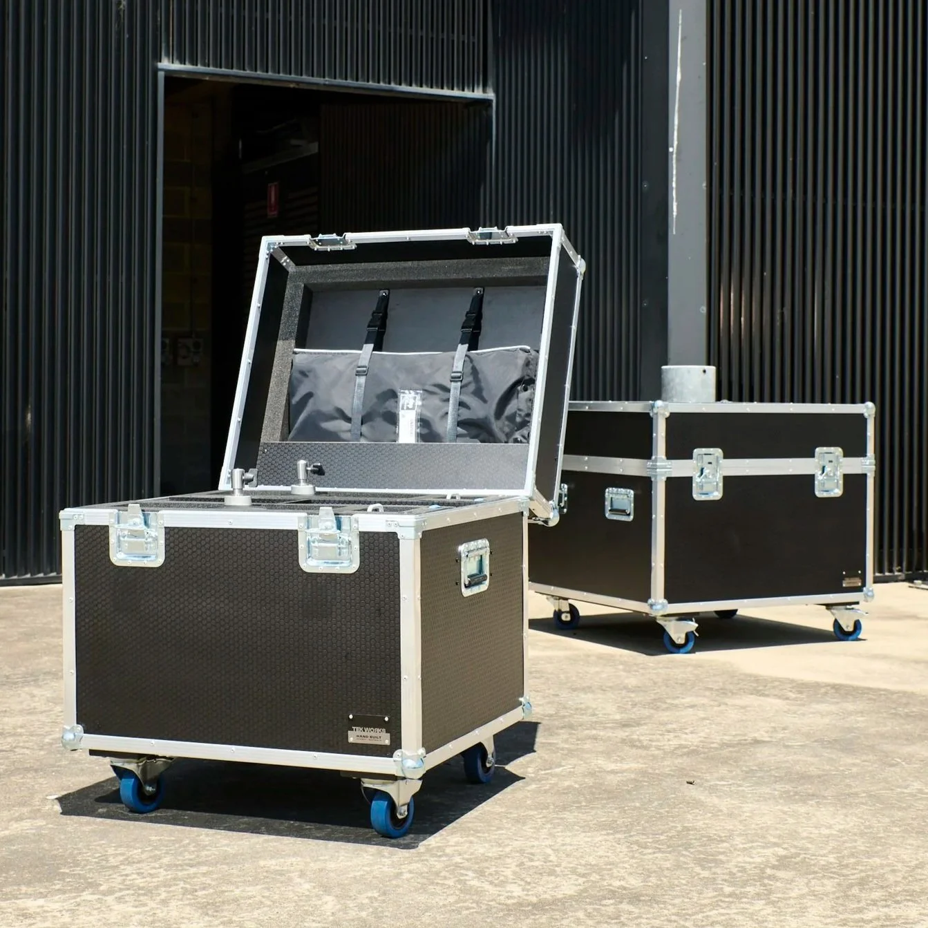 Two large black and silver equipment cases with wheels, one of which is open showing foam interior and straps, situated outside against a black metal building.